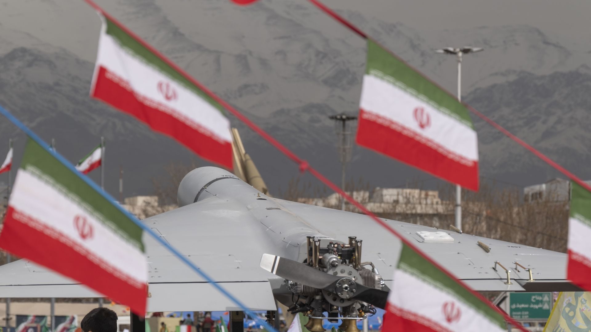 Crowd at a festival with Iranian flags strung overhead; a gray helicopter engine and propeller sit in the foreground, mountains visible in the background.