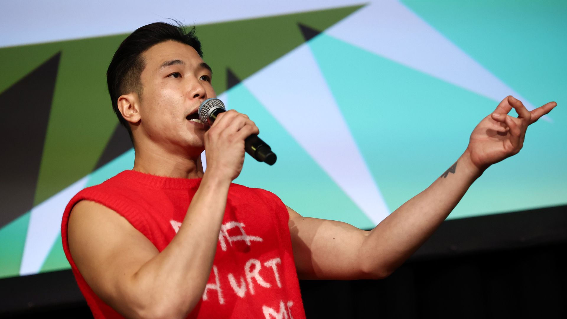 Photo of Joel Kim Booster speaking with a microphone while gesturing with his left arm