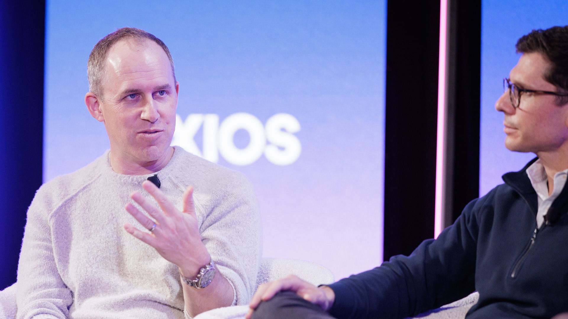 Two men seated on white armchairs in a panel discussion. One man in a light gray sweater gestures while speaking, the other in glasses and a navy zip-up listens. Blue background with AXIOS logo.