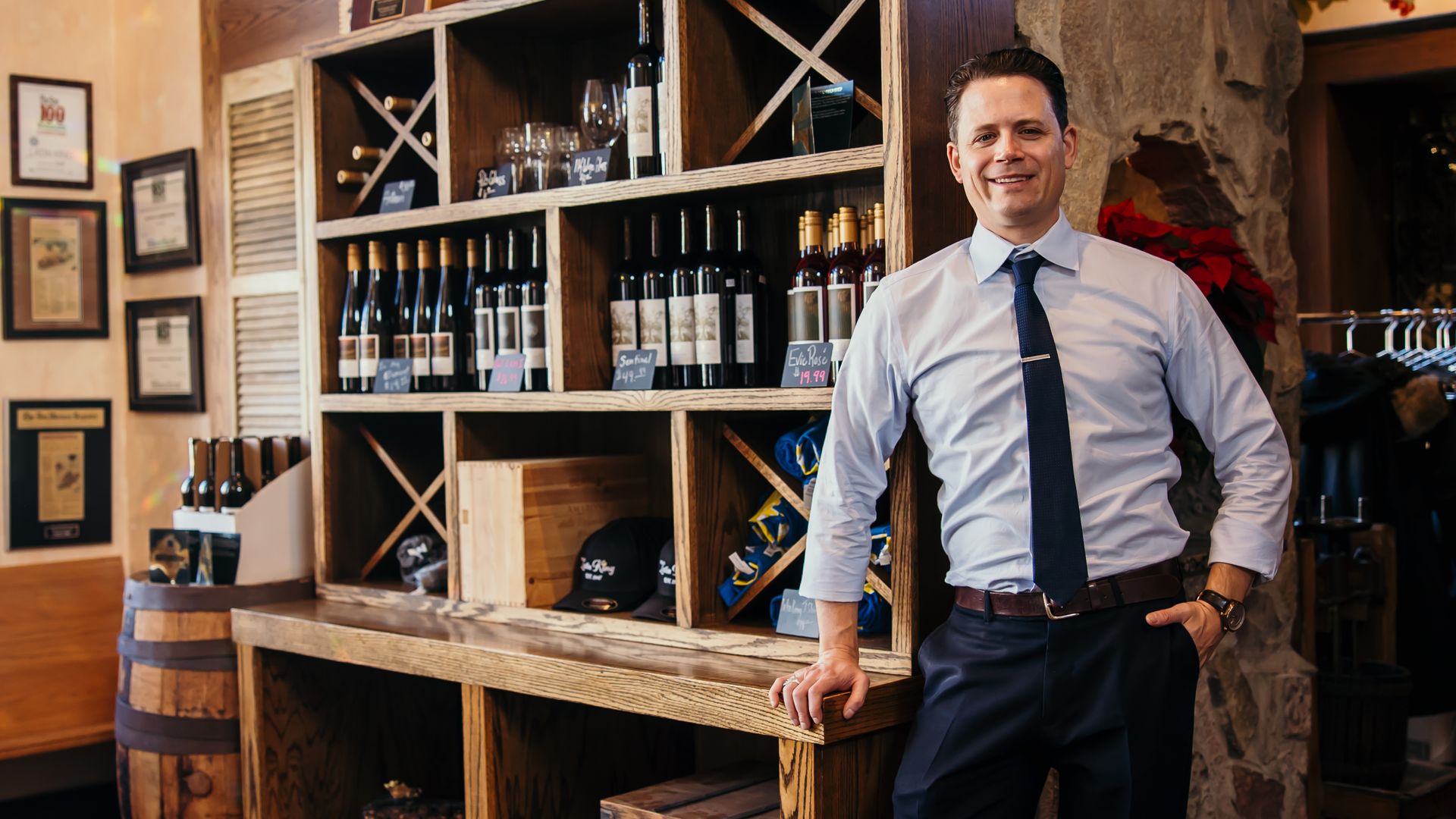 Man in white shirt and dark tie standing next to wooden shelf filled with wine bottles and glassware inside rustic store with stone wall and barrel.