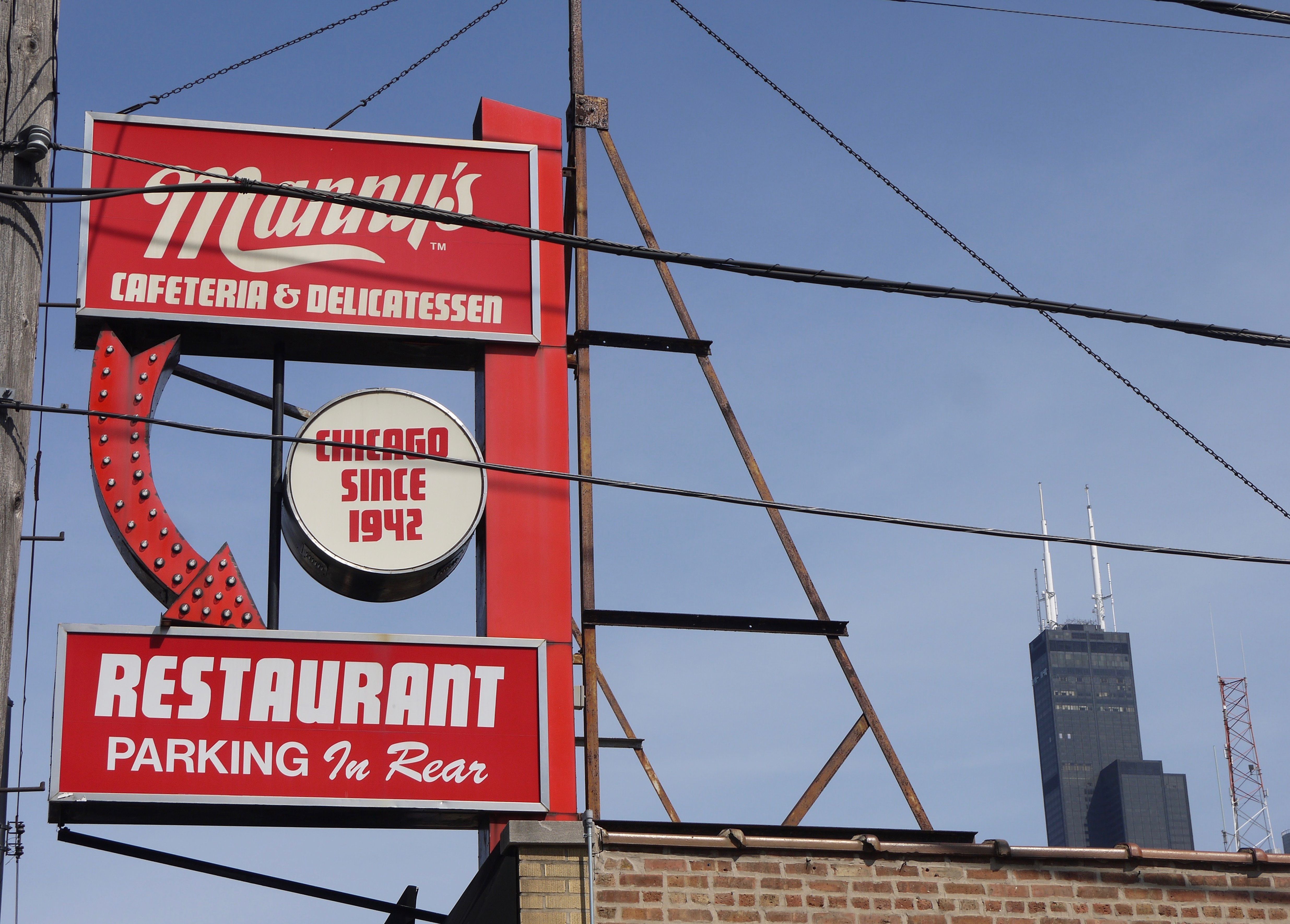 Red restaurant sign with white cursive logo and "CAFETERIA & DELICATESSEN", mounted on a metal frame. A circular sign below reads "CHICAGO SINCE 1942"; blue sky in the background.