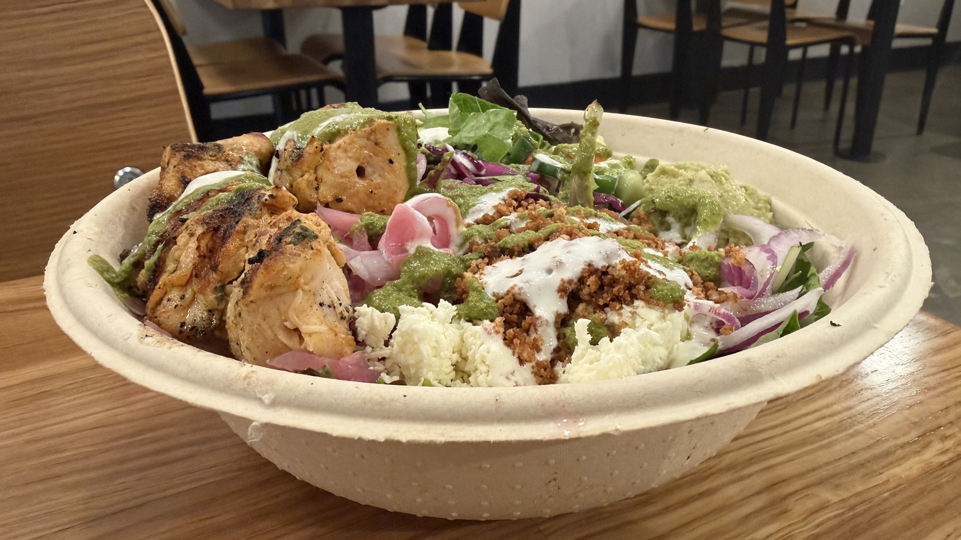 A large disposable bowl holds a Mediterranean salad: grilled chicken chunks, feta crumbles, red onions, cucumber, mixed greens, and green dressing, on a table in a casual dining area.