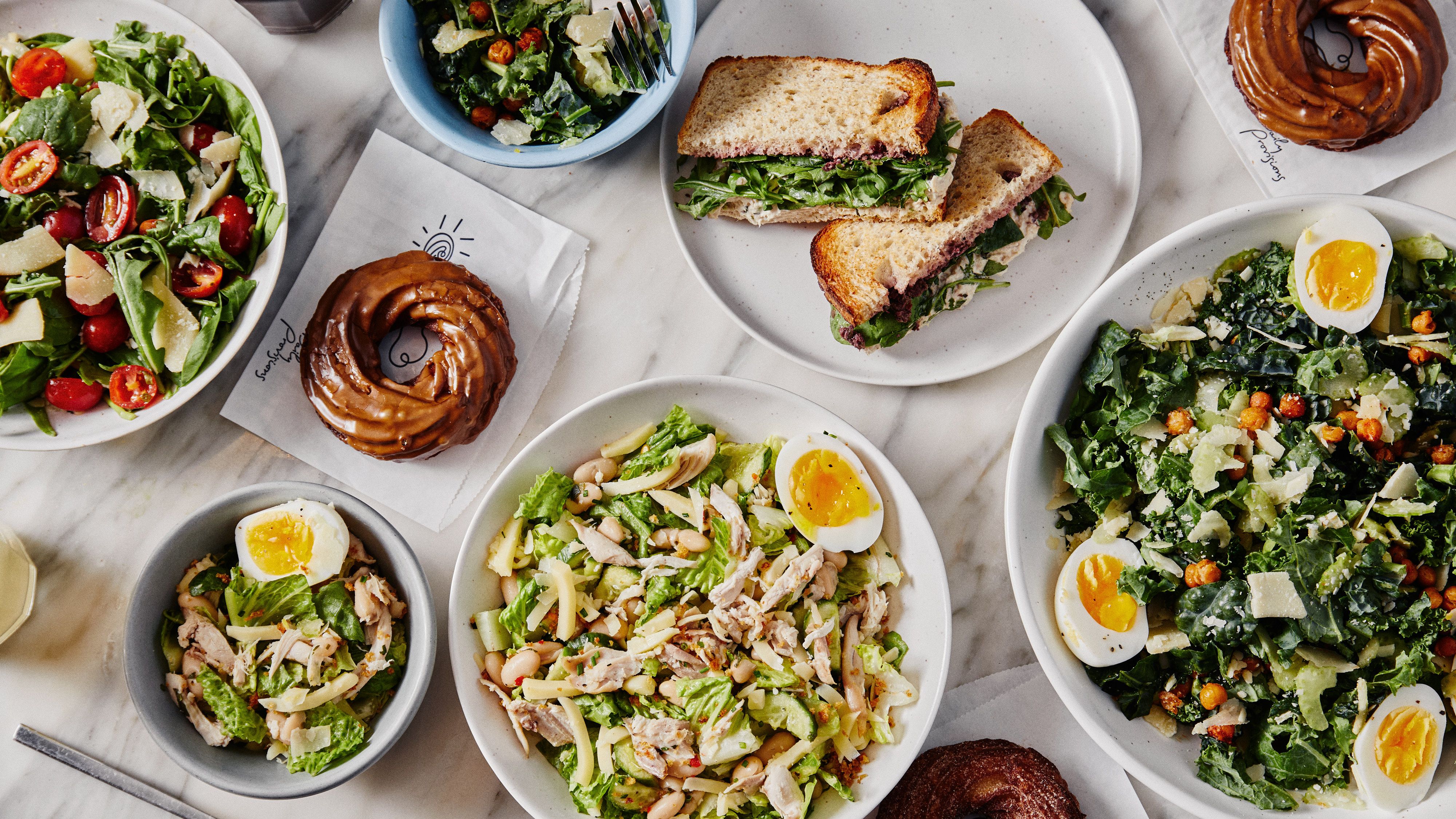 A variety of salads with greens, cherry tomatoes, hard-boiled eggs, shredded chicken, and cheese, two chocolate-glazed donuts, and a sandwich with leafy greens on a white table.