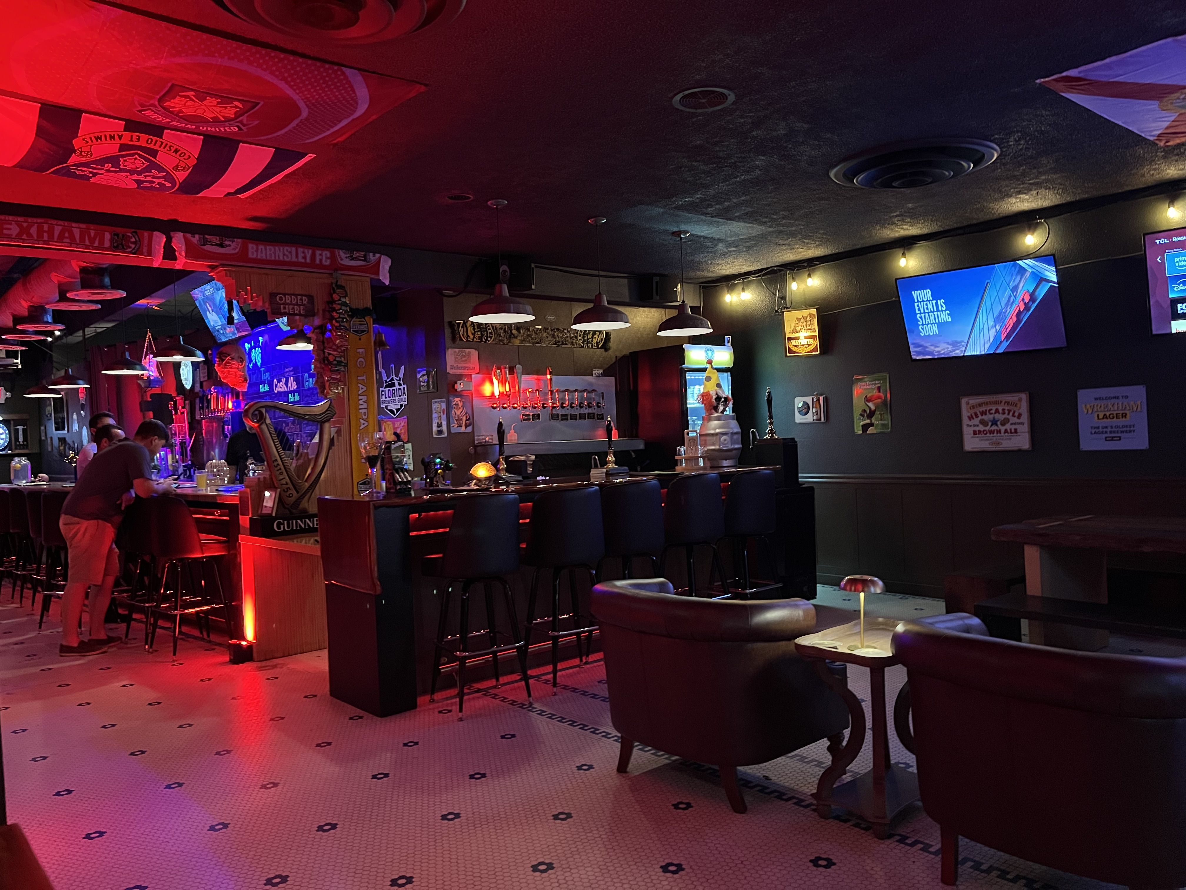 Dimly lit bar with red and blue lighting, black bar stools, leather chairs around a small table, sports flags and signs, and TVs showing sports content on dark walls.