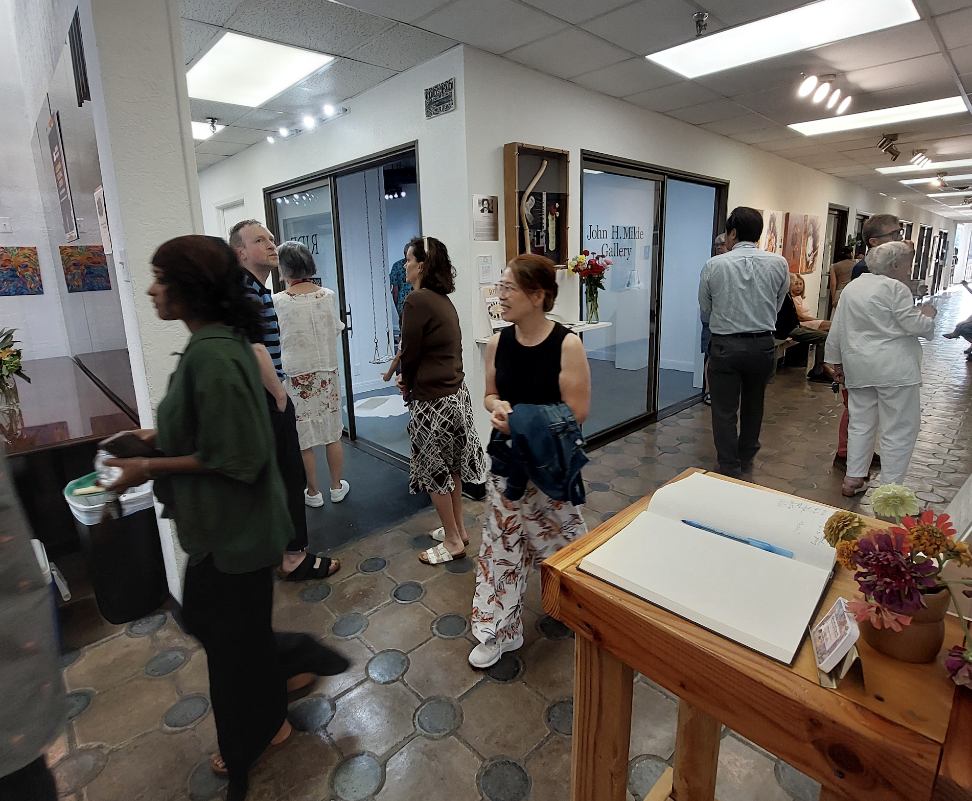 People gather in a tiled hallway outside the John H. Milde Gallery in the Goldmark Cultural Center. Some are looking at art, others walking, with a wooden table holding an open guestbook and flowers in the foreground.