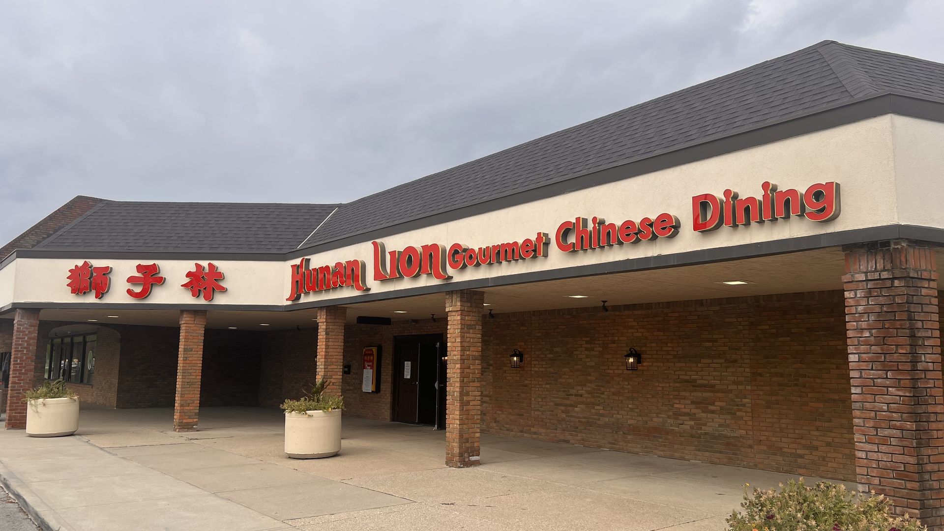 Exterior of Hunan Lion Gourmet Chinese Dining restaurant with red signage, brick pillars, and planters with flowers under cloudy sky.