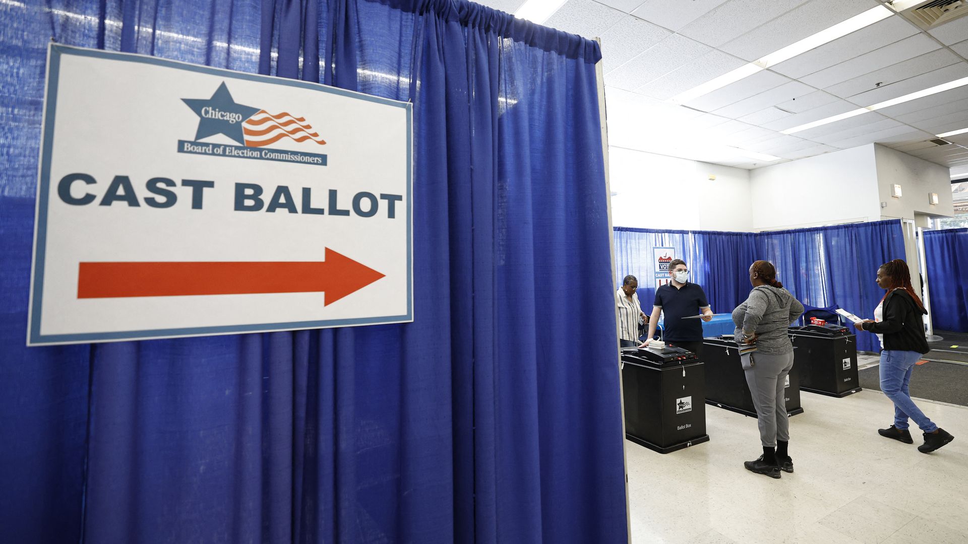 Chicago voting site with blue curtains and a large "CAST BALLOT" sign with red arrow, people lined up to cast ballots in black ballot boxes inside the bright room.