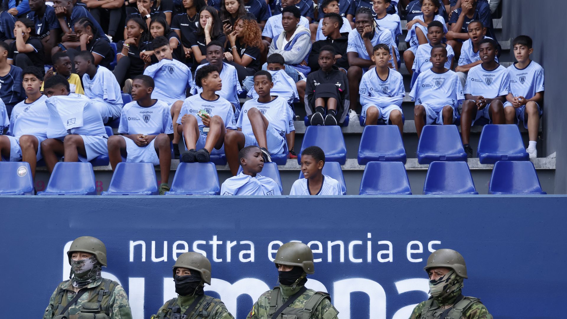 Four military officers dressed fully in fatigues stand below rows of young soccer players in blue jerseys at a futbol stadium