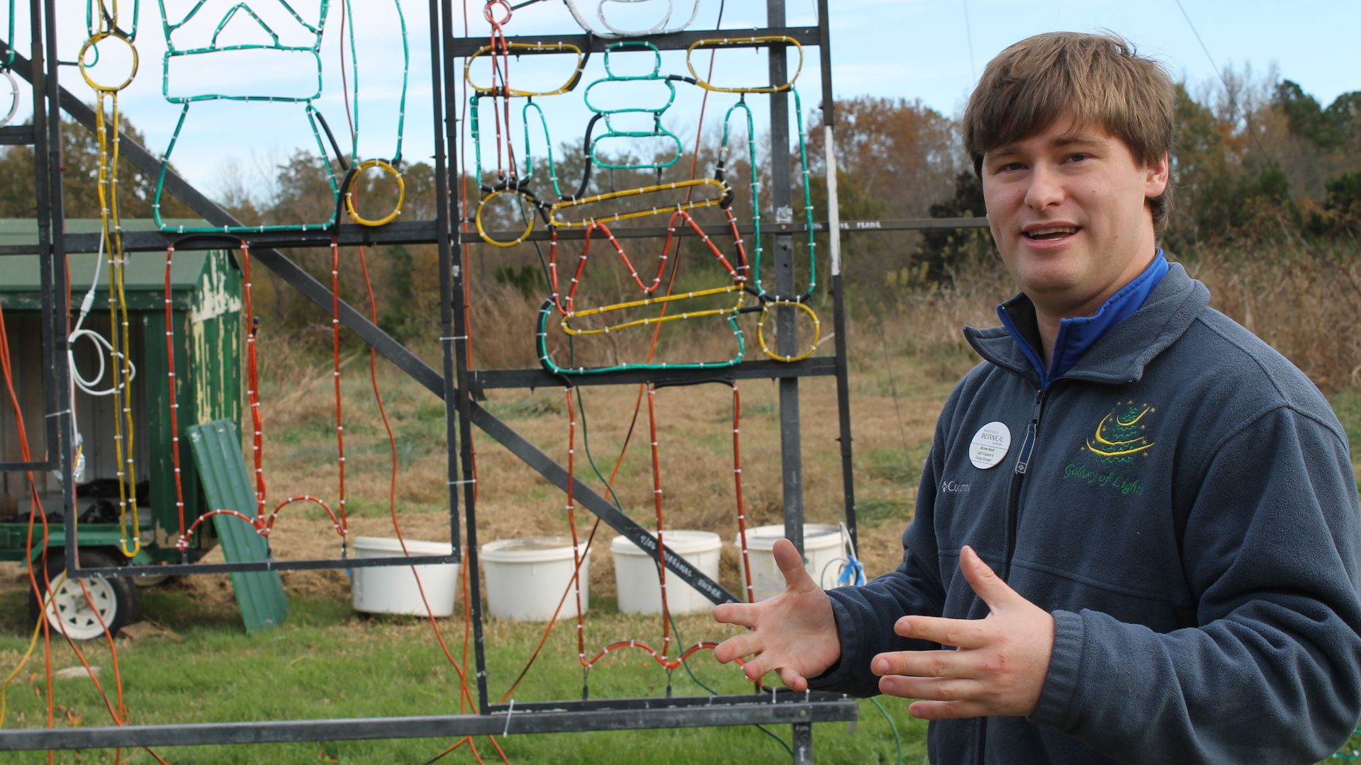 Man in dark blue fleece jacket with a name tag explaining outdoor light display shapes on metal frame, grassy field and trees in background under partly cloudy sky.