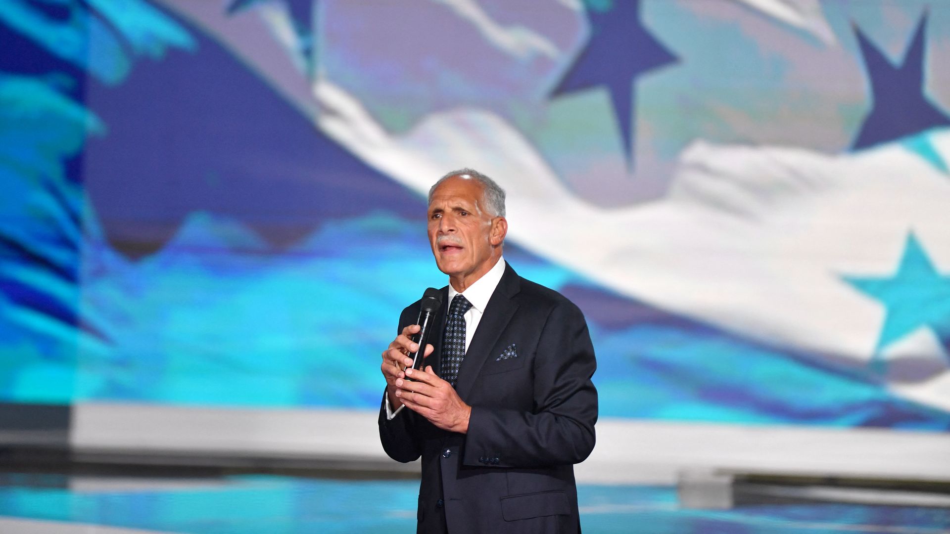 A gray-haired, balding and thinly moustachioed Honduran presidential candidate and businessman Nasry Asfura, wearing a navy suit jacket, white shirt and black tie with gray checks, clasps a black mic as he speaks onstage at an event in front of Honduras' blue and white flag, with four stars.