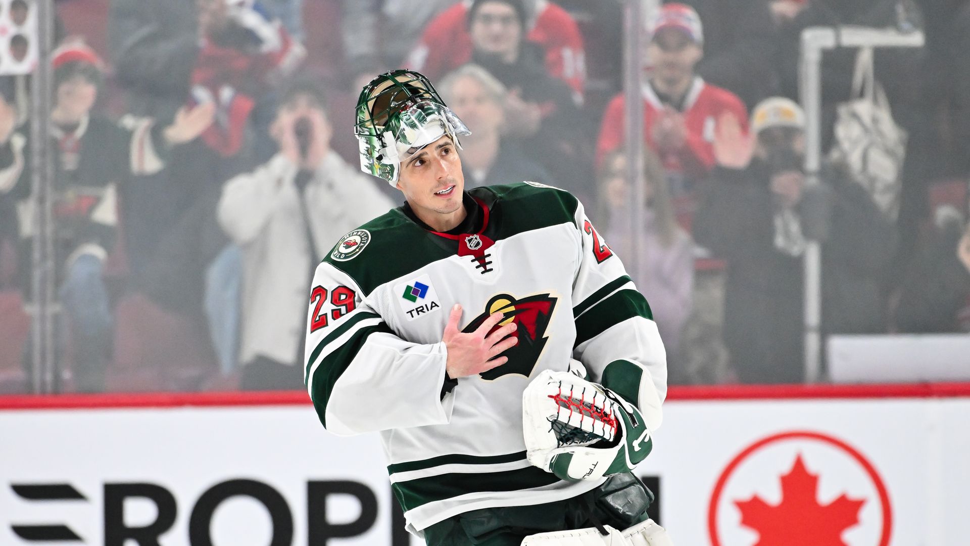 A goaltender with his helmet flipped up and his face visible holds his hand to his heart on the ice as the crowd in the background applauds