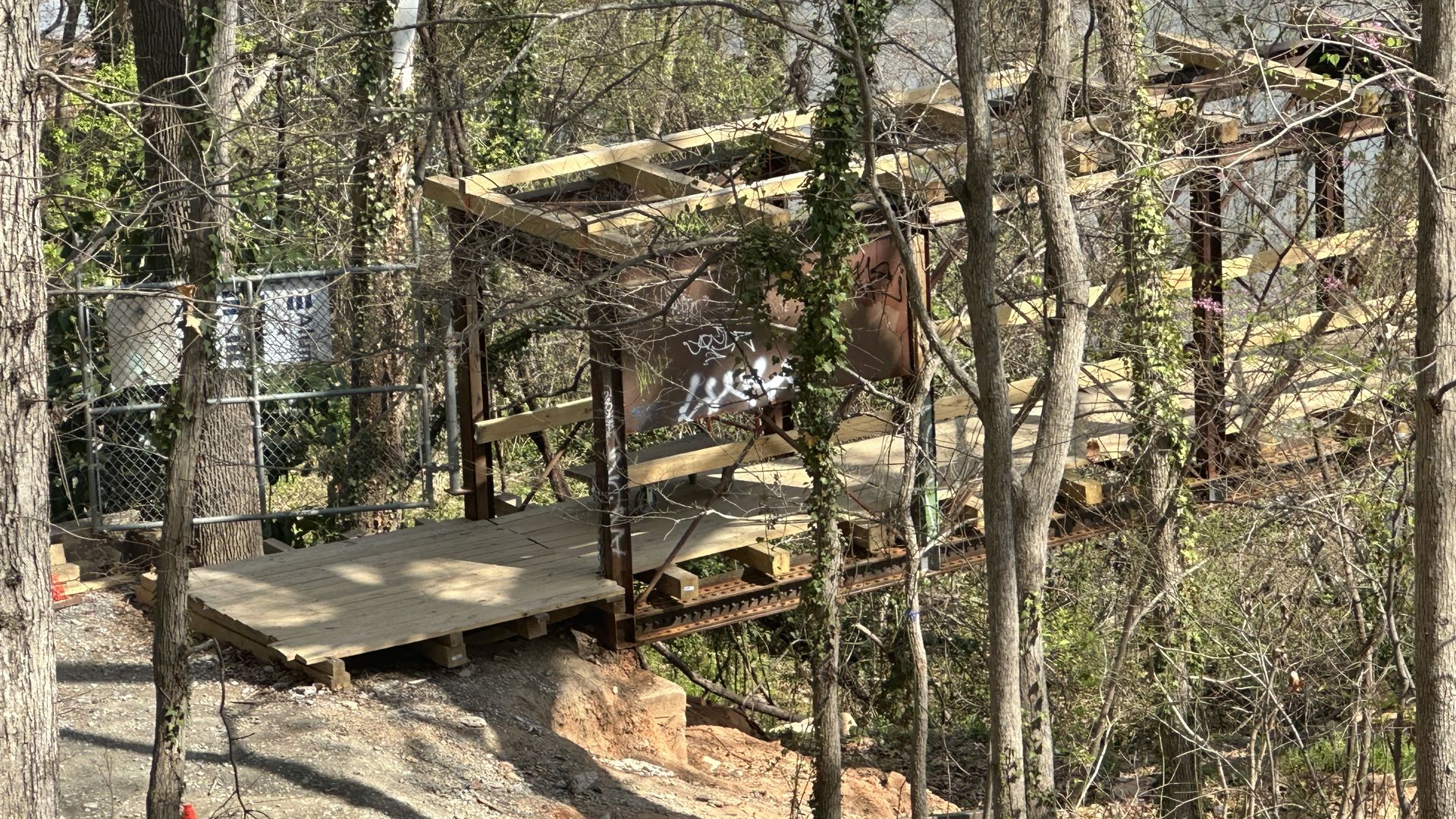 Forest scene with a partially built wooden elevated platform and railing, stairs on the right; a chain-link fence to the left; a light-brown dog on the rocky, leaf-strewn ground.