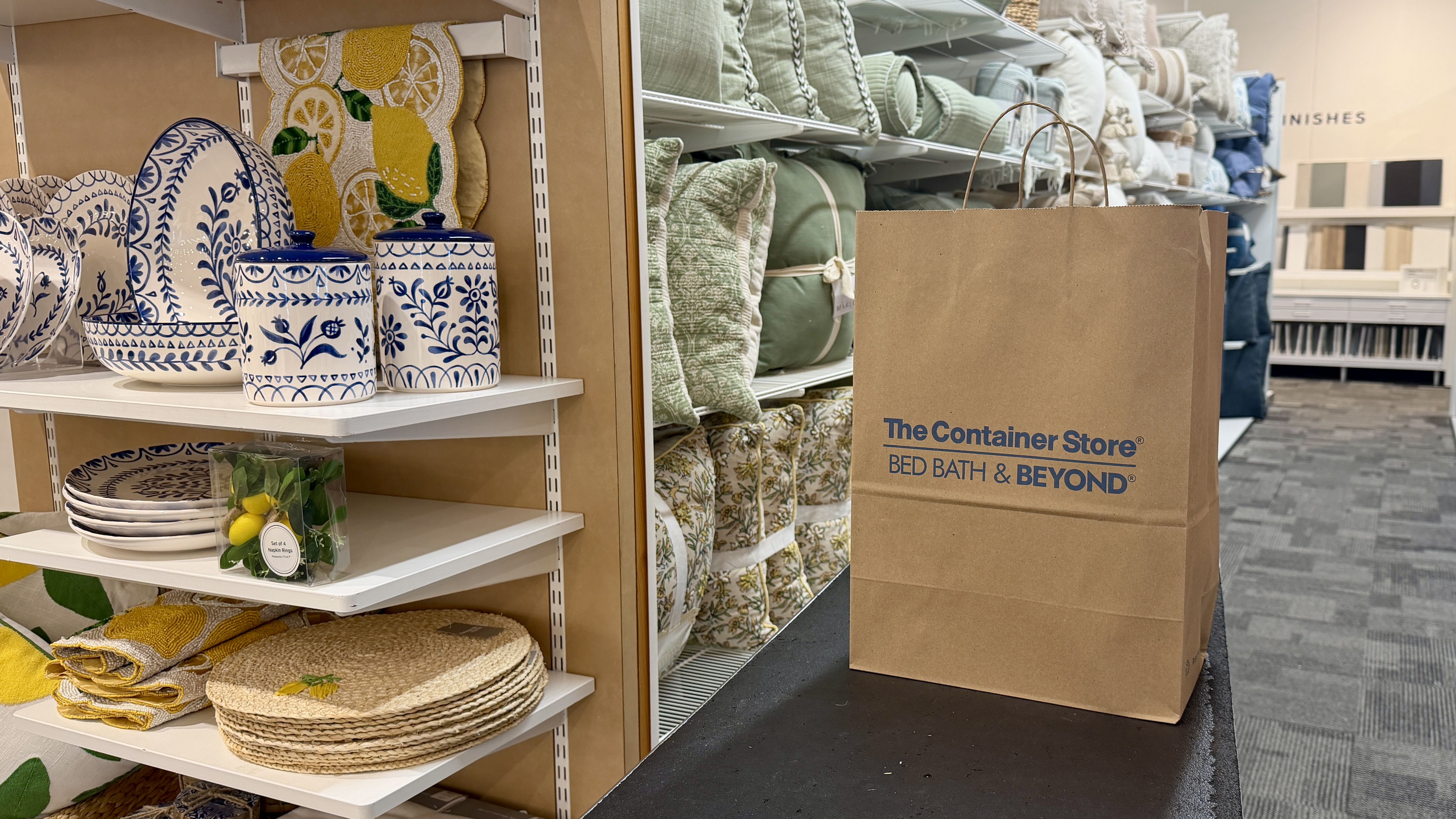 Home goods aisle with blue-and-white ceramic canisters and plates, woven placemats, and lemon textiles; a large brown The Container Store bag sits on the right with folded textiles in the background.