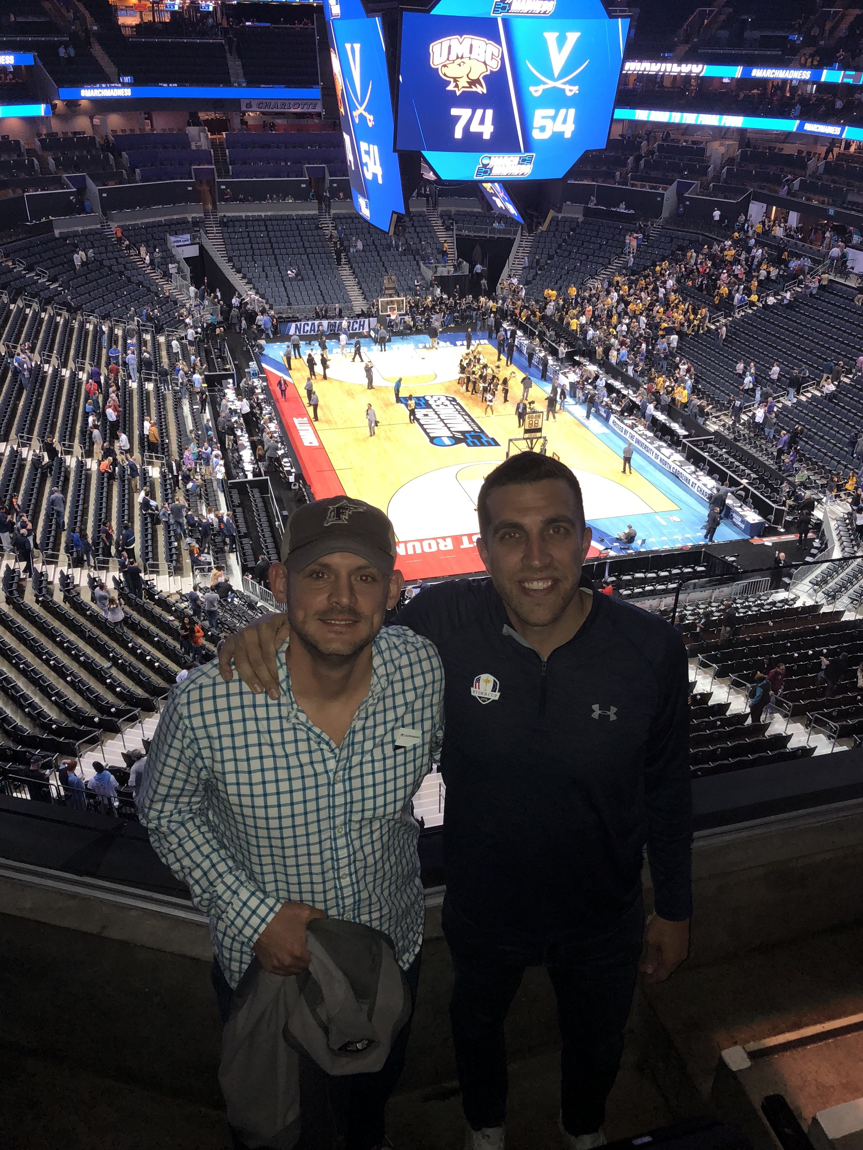 Two men smiling in a stadium with a basketball court below showing UMBC leading 74-54 against Virginia on the scoreboard during an NCAA March Madness game.
