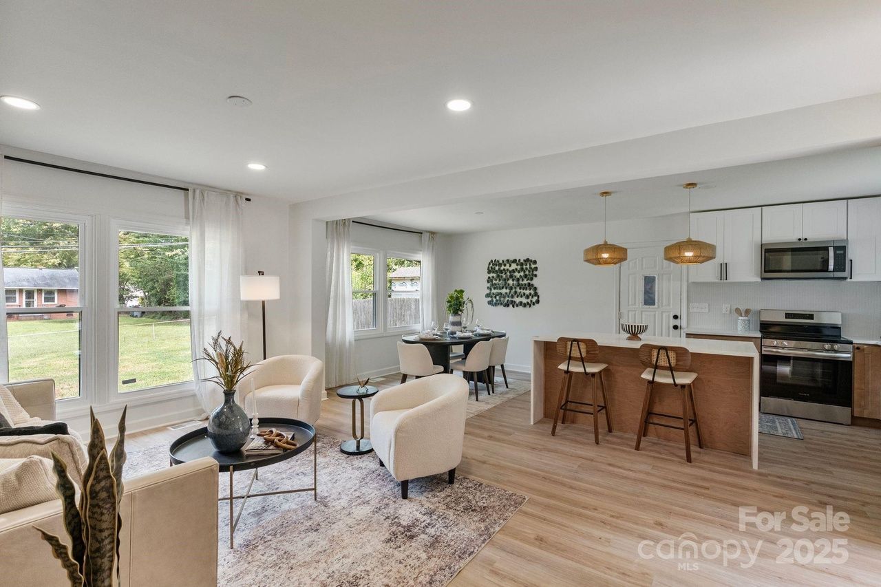 Bright open-concept living space with beige armchairs, round black table, dining area with black chairs, wooden kitchen island with two stools, white cabinets, and large windows.