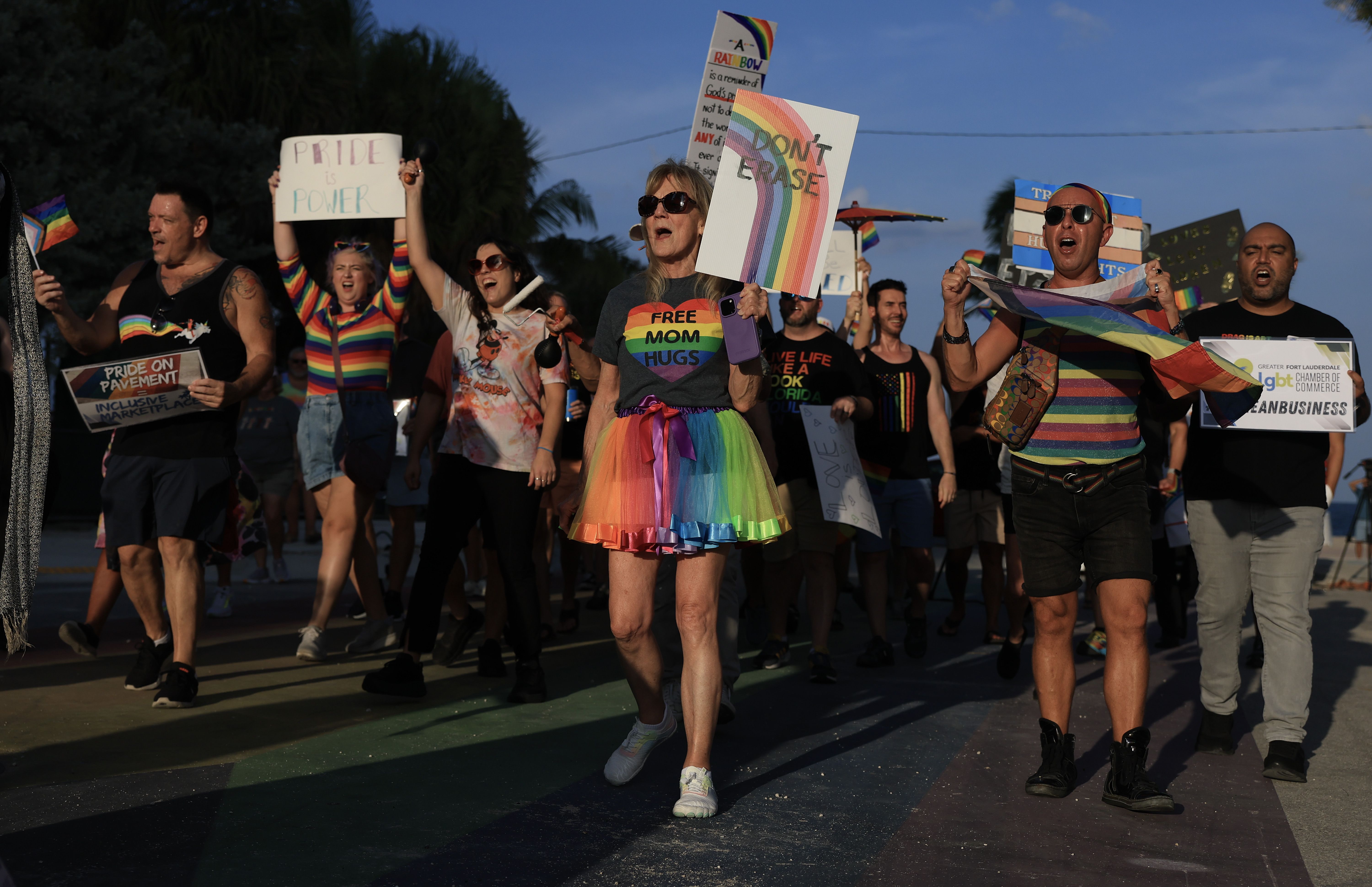 Group of diverse people marching at a pride event carrying rainbow flags and signs, including "Don't erase" and "Pride is power," wearing colorful rainbow-themed clothing.