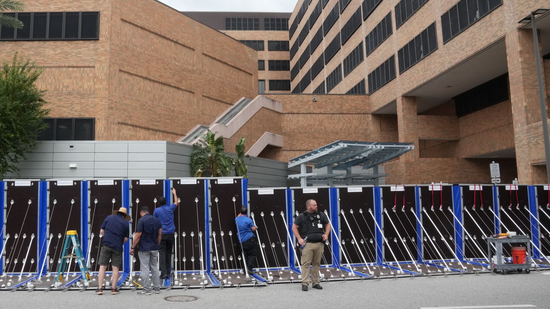 Five men working on or near a long black and blue AquaFence flood barrier set up on a street in front of beige brick buildings under a gray, cloudy sky.