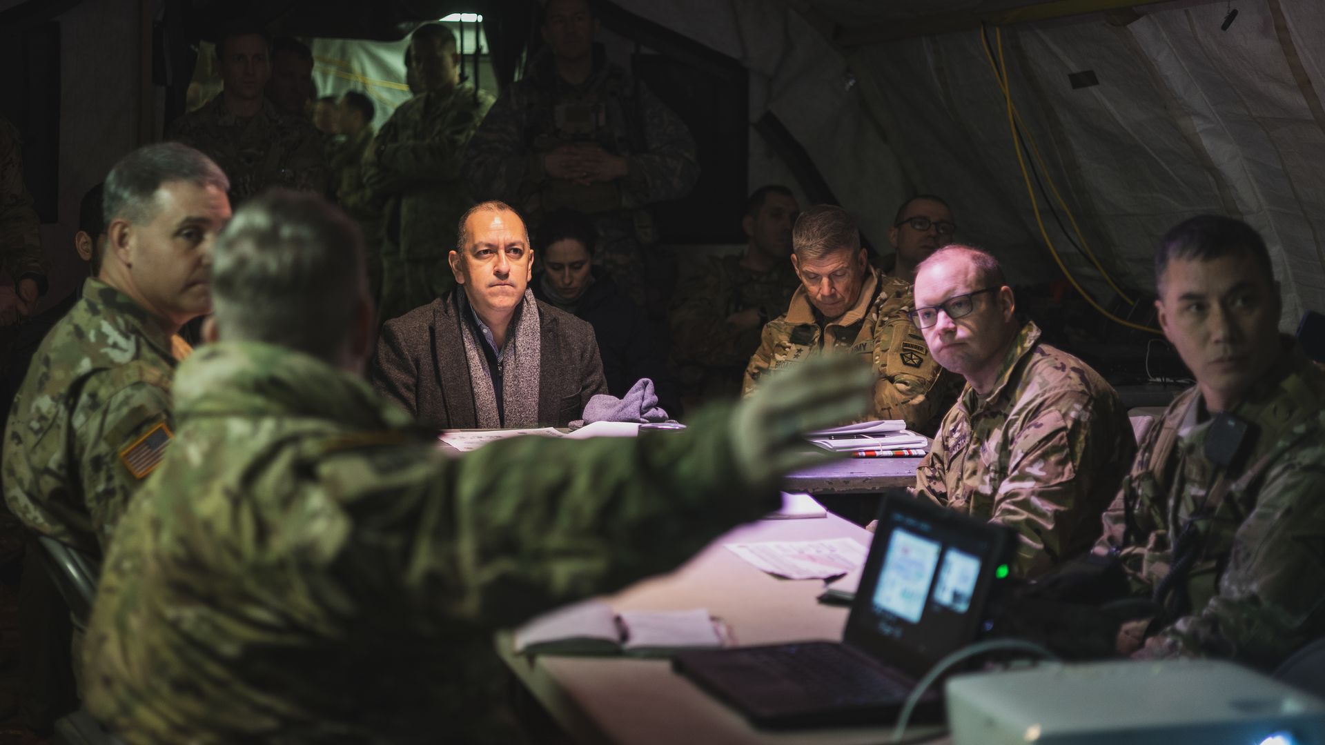 Men sit around a table in a dark tent. At the head of the table is Gabe Camarillo, the Army undersecretary. He is in a black coat and charcoal scarf.