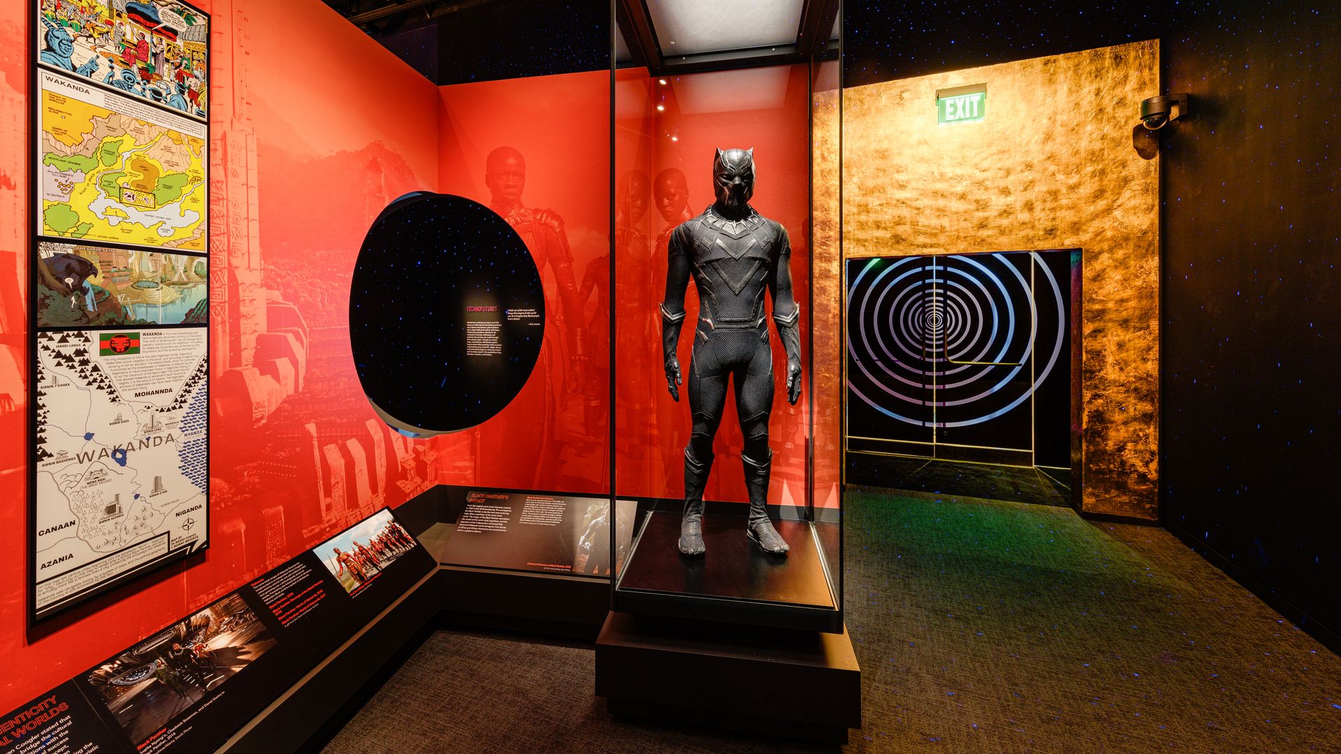 A museum exhibit with a mostly red and orange background. In the foreground is a superhero suit wore by Black Panther in a glass case.