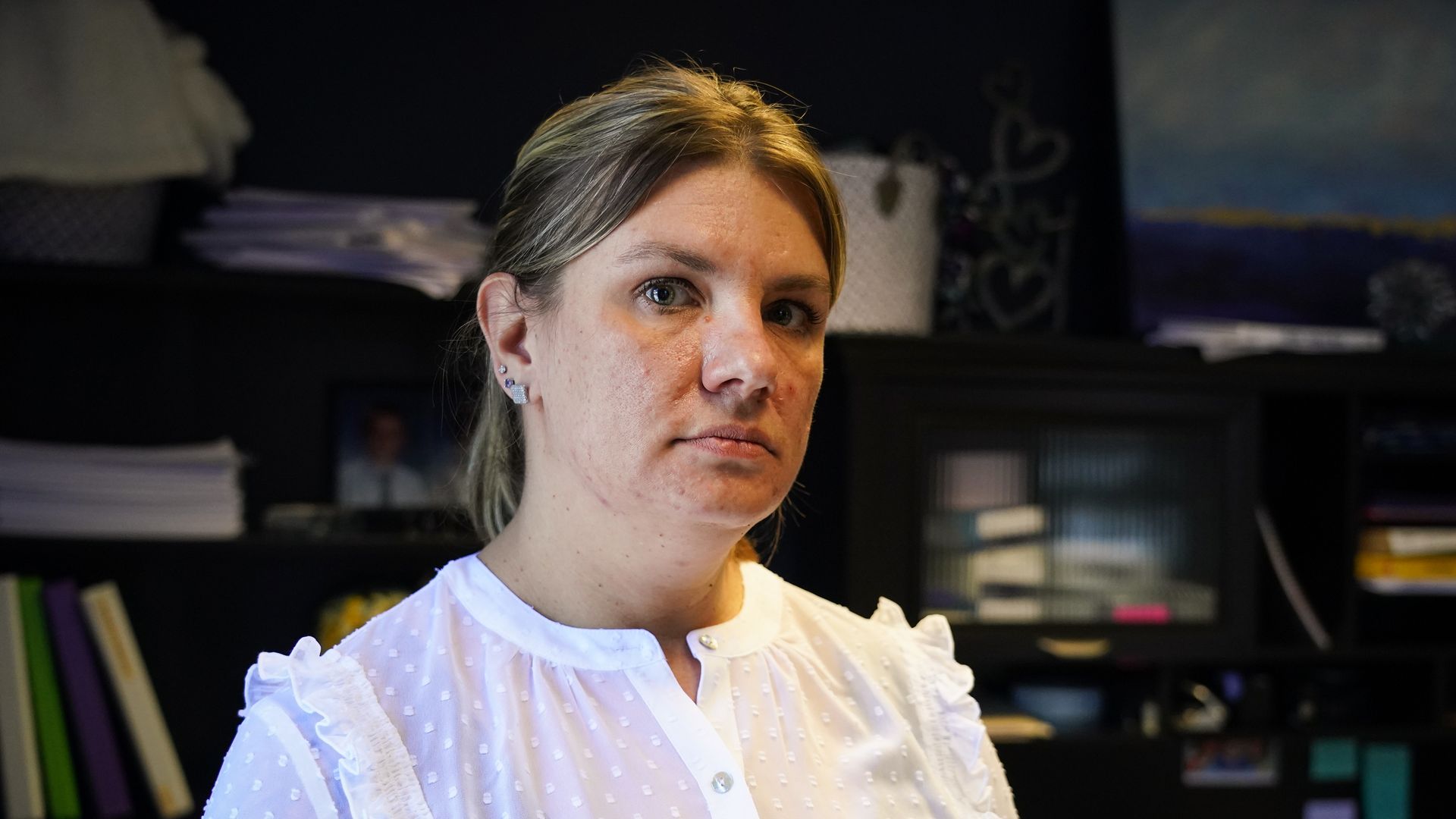 Portrait of a woman with blonde hair and blue eyes, wearing a white dotted blouse, standing indoors with dark shelves and stacks of papers behind her.