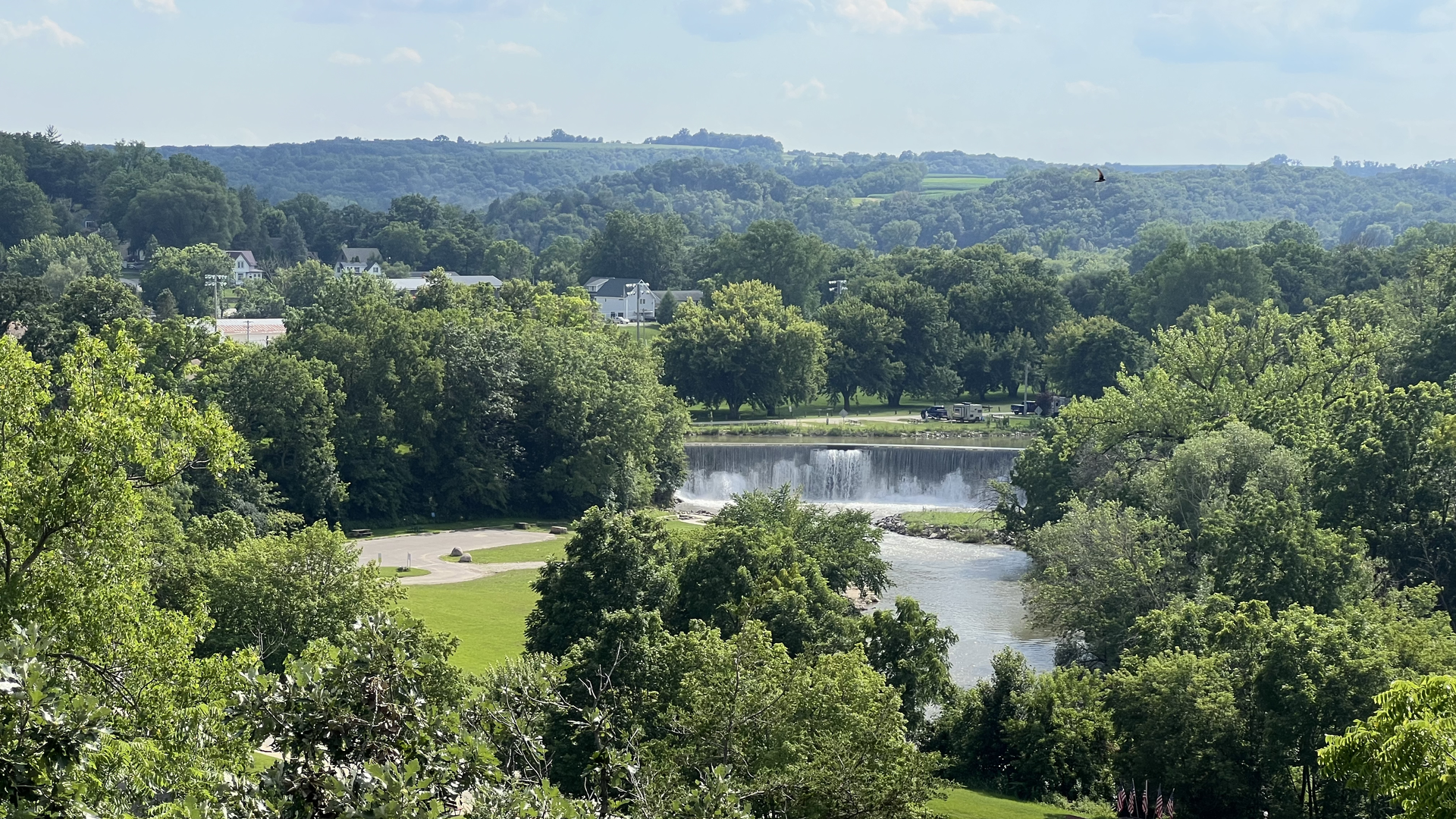 A rolling landscape populated by lush green trees, a blue sky and a dammed river in the foreground