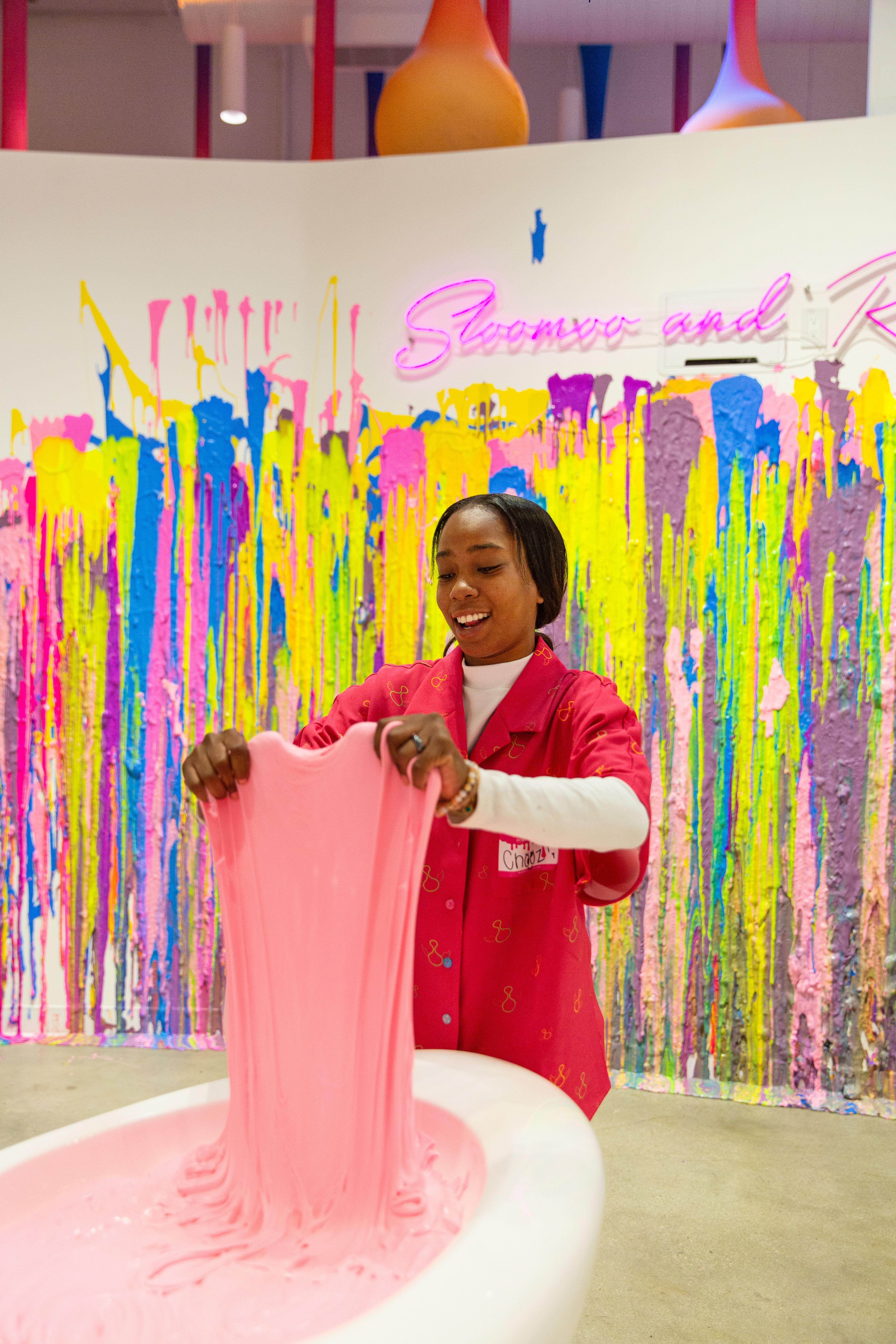 Woman in a red shirt stretching pink slime over a white tub, with a colorful paint-splattered wall and a neon sign behind her.