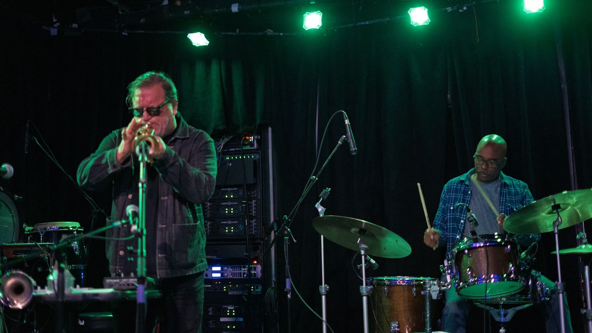 Dim stage with green spotlights; a trumpet player in sunglasses and dark jacket on the left, and a drummer in a plaid shirt on the right, with drums and amplifiers in the background.