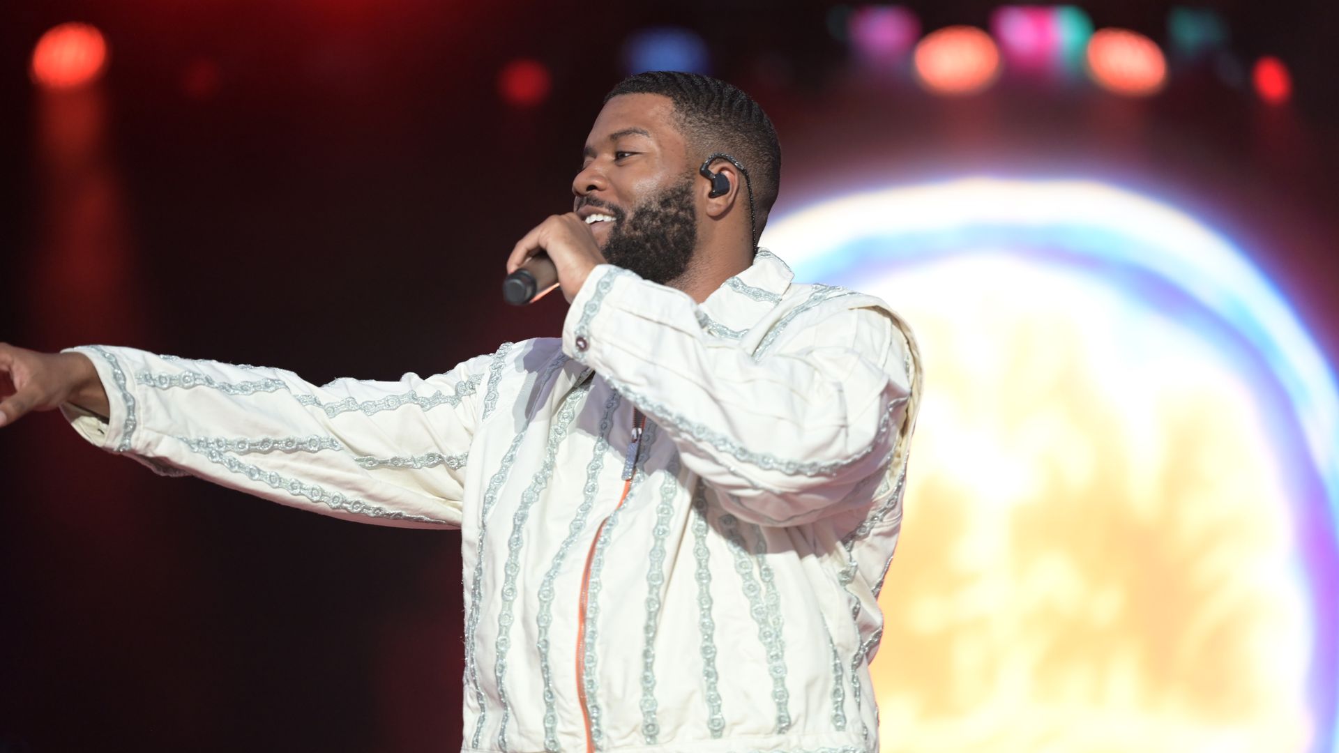 A male singer on stage, wearing a white embroidered shirt and an in-ear monitor, holding a microphone near his mouth with his right hand as his left arm extends. Bright, blurred stage lights in the background.