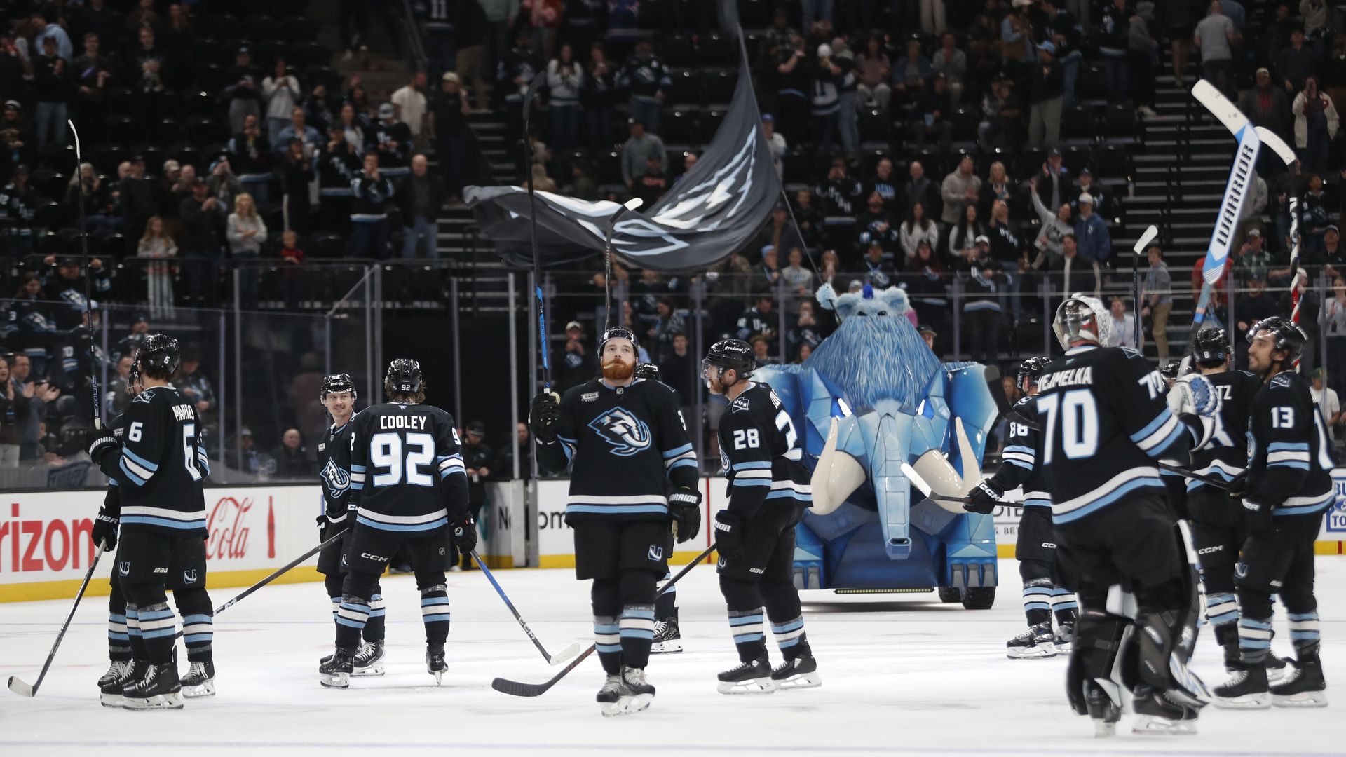 Ice hockey scene with players in black and light-blue jerseys, including #92 Cooley and #70; a blue winged mascot stands near the center as the cheering crowd fills the arena.