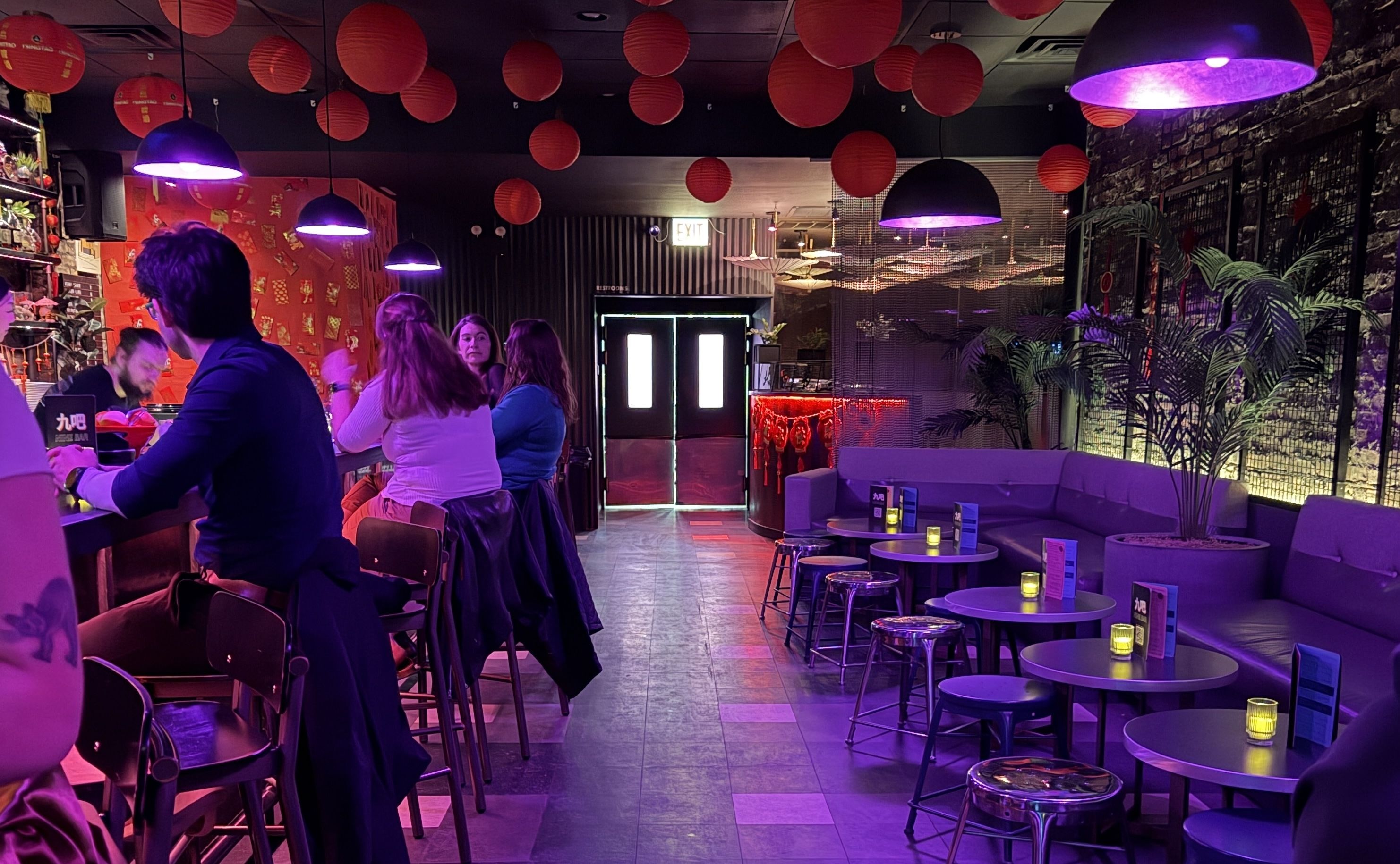 Dimly lit bar with purple lighting, red lanterns hanging from ceiling, people seated at the bar, small tables with stools and candles on the right, plants by a brick wall.