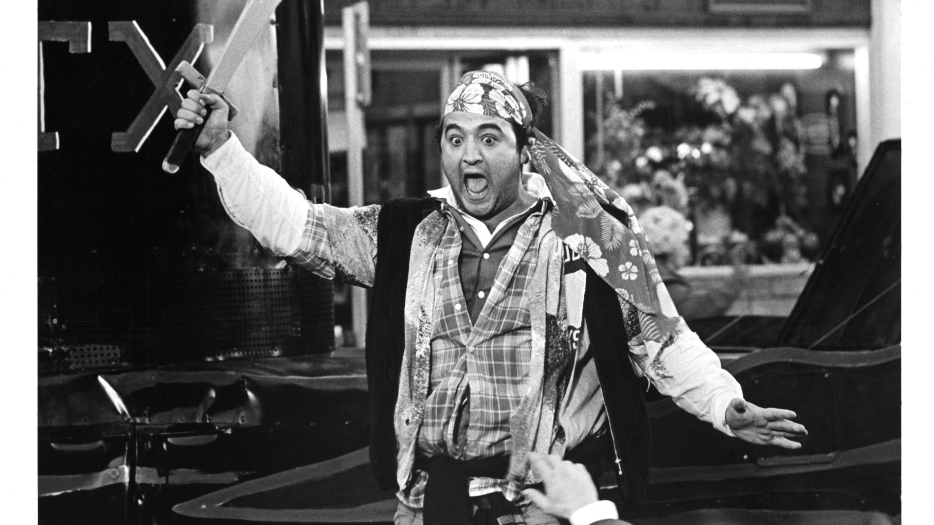 Black and white photo of a surprised man wearing a floral headscarf and layered clothing, holding a curved sword above his head with his mouth open, standing near a vehicle.