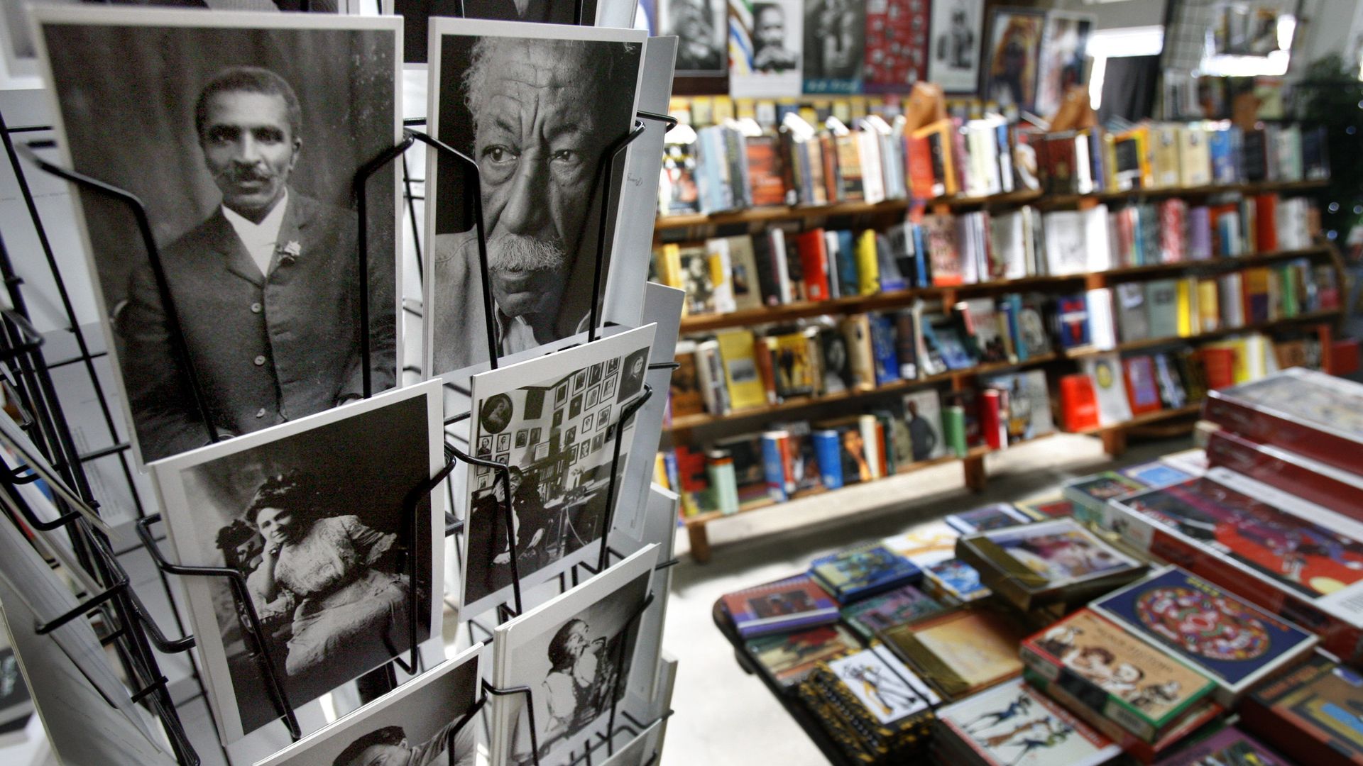 Black-and-white portrait cards on a rotating rack in a bookstore, featuring historic figures; shelves of colorful books line the background, with tables of stacked books in the foreground.