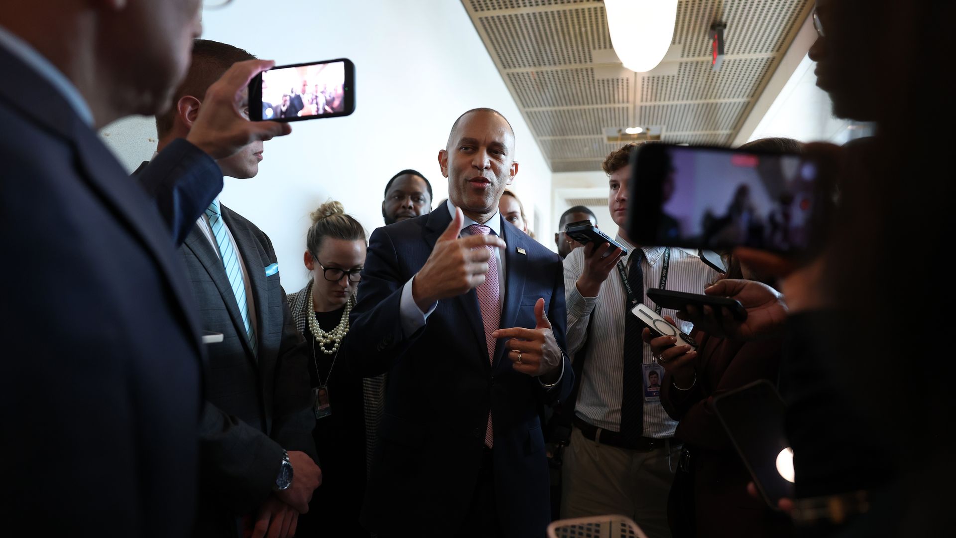 House Minority Leader Hakeem Jeffries speaks to reporters in a white-halled basement while flanked by staff and security.