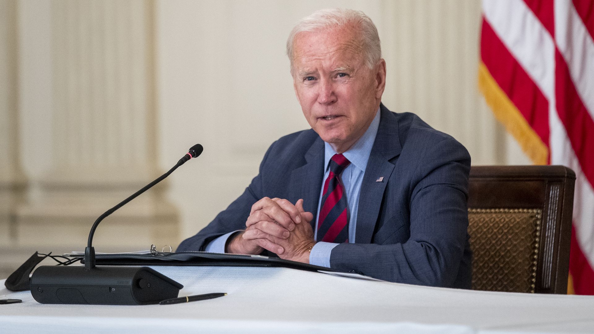 President Joe Biden sits at a meeting with a microphone in front of him while his hands are clasped together