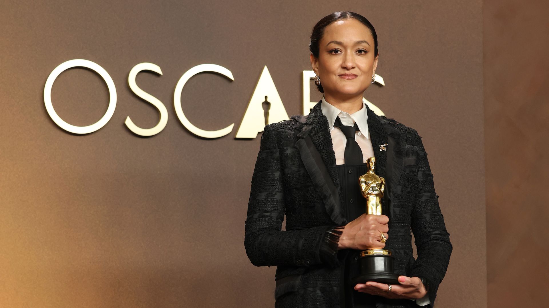 A poised woman in a black textured suit with a white shirt and black tie holds a gold Oscar statuette on stage, with large OSCARS lettering behind her and warm amber lighting.