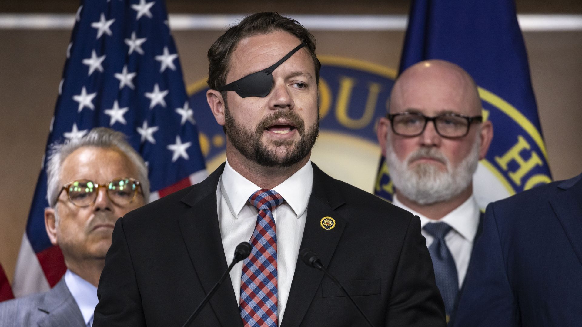Man with black eye patch, beard, and black suit with checkered red and blue tie speaks at a podium, flanked by two men in suits and glasses, with American flags in background.
