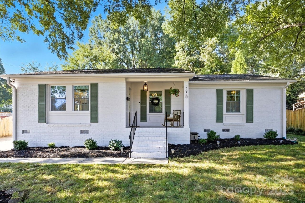 White brick single-story house with green shutters and front porch featuring a hanging plant and chair, surrounded by trees and a lawn under a clear blue sky.