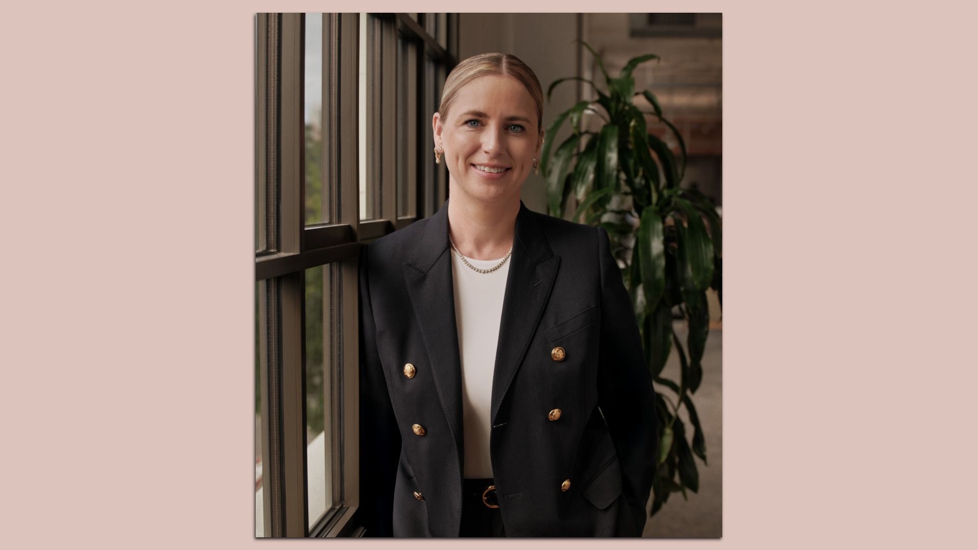 Portrait of a smiling blonde woman in a black double-breasted blazer with gold buttons, white top, and pearl necklace, standing by a window with a green plant in the background.
