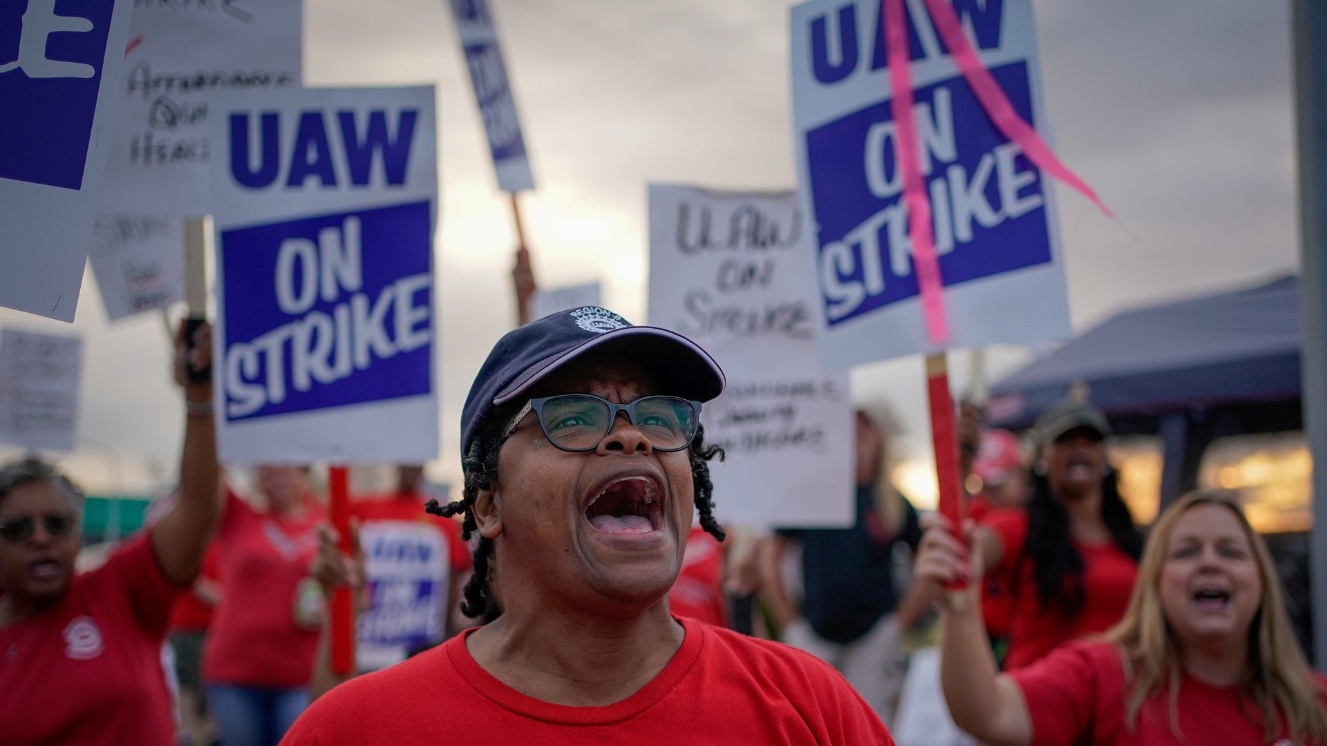 In this image, a woman shouts while standing in front of people holding signs that say "UAW strike" 