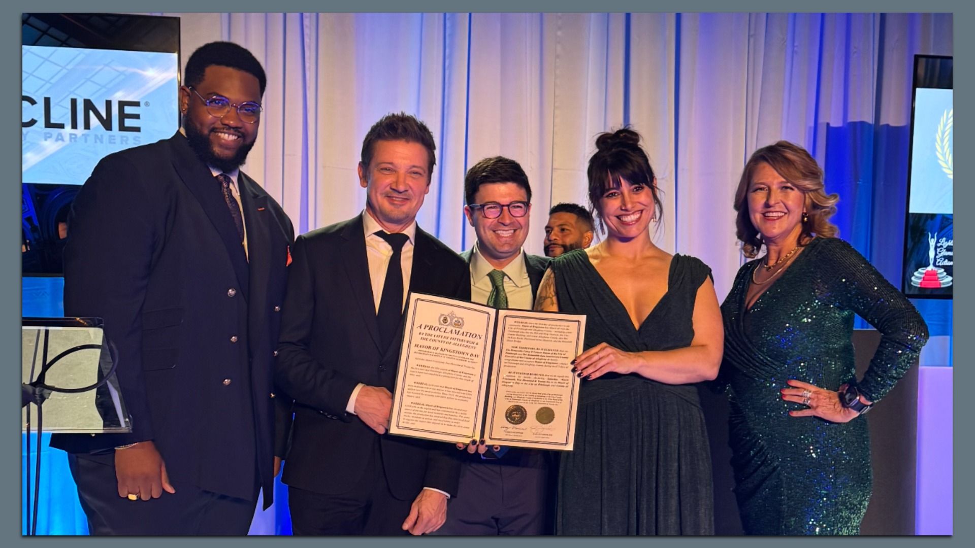 Five formally dressed people on a stage smile at the camera while holding an open certificate; blue-lit curtains and a screen in the background indicate a formal awards event.