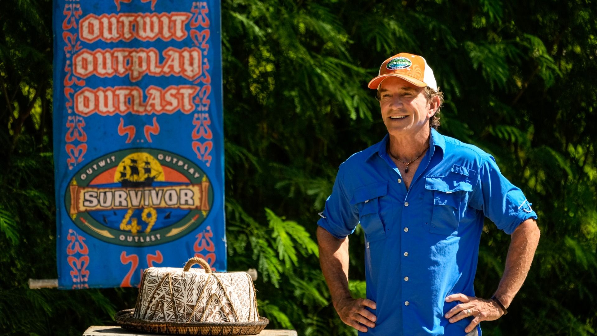 "Survivor" host Jeff Probst stands with his hands on his hips during Season 49 in a blue shirt and orange hat smiling with a sign to his left saying "OUTWIT OUTPLAY OUTLAST" and "SURVIVOR 49."