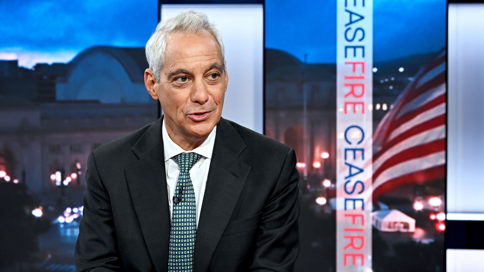 Rahm Emanuel in suit and green tie in front of a green screen with CEASEFIRE in blue and red lettering and an American flag/