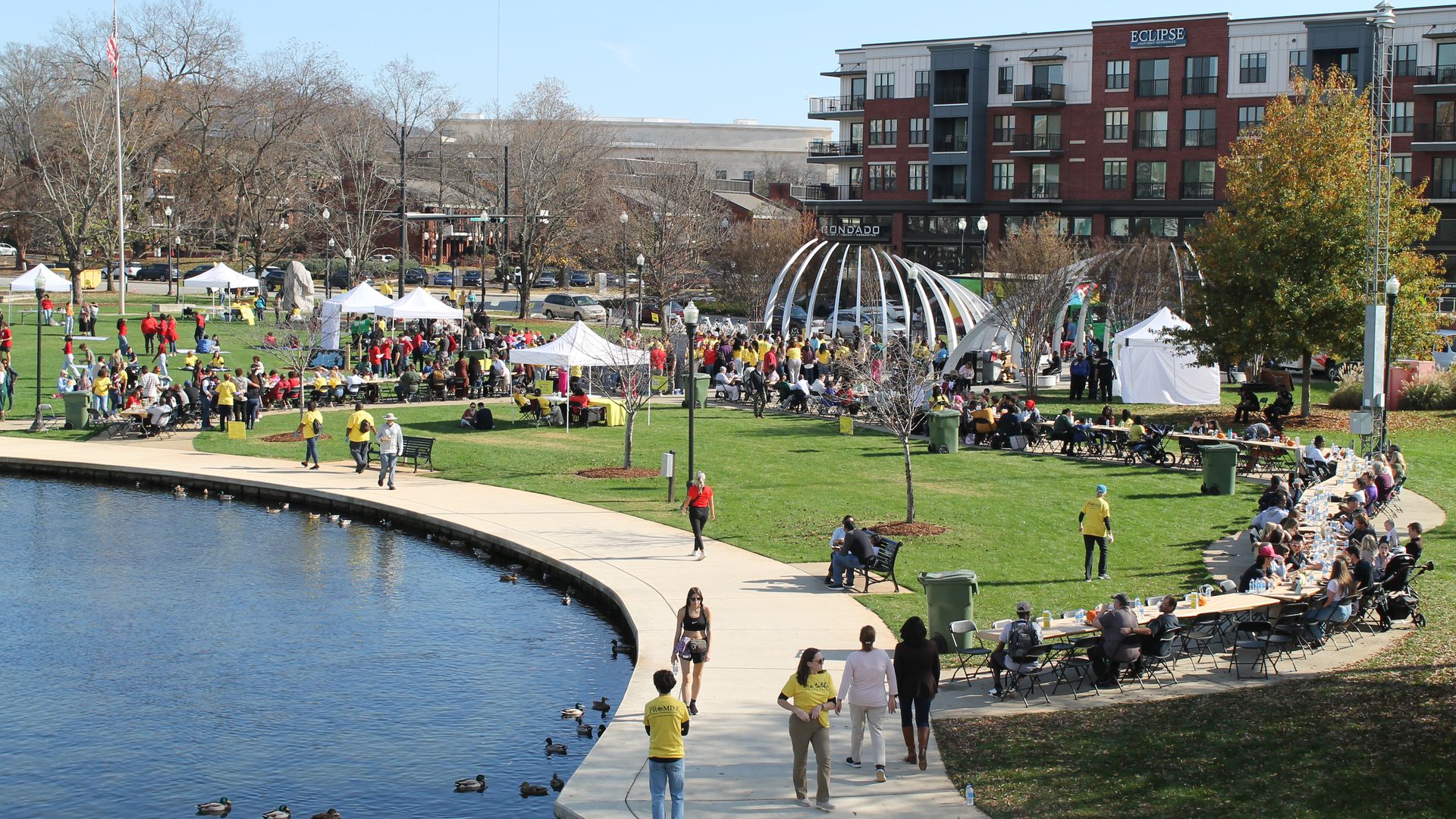 Outdoor community event by a pond with people gathered under white tents, sitting at long tables on green grass near modern red brick apartment buildings under a clear blue sky.