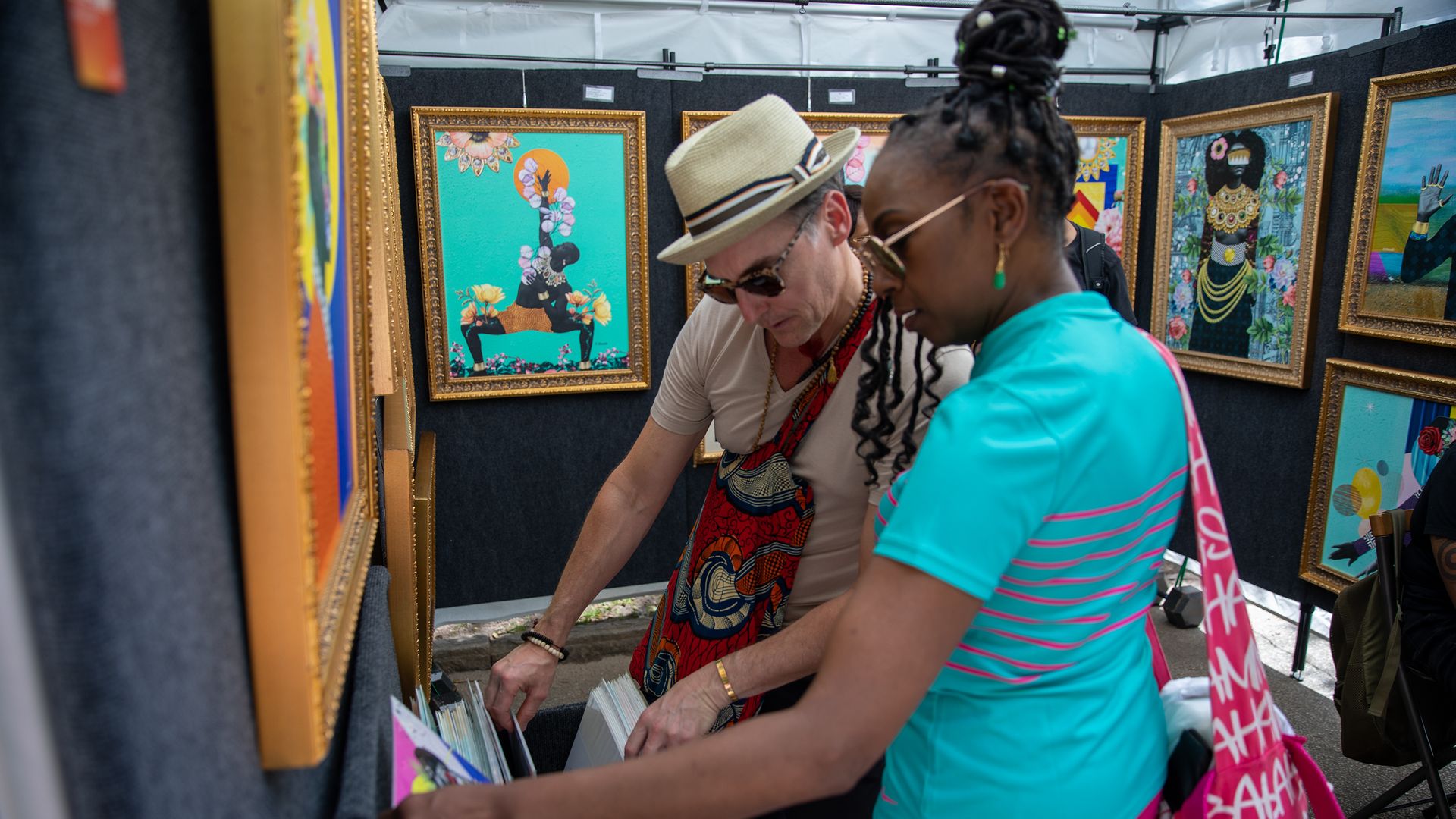 Two art shoppers browse a stall of framed colorful paintings; a man in a beige shirt and straw hat leans over vinyl records while a woman in turquoise top and pink pants stands beside him.