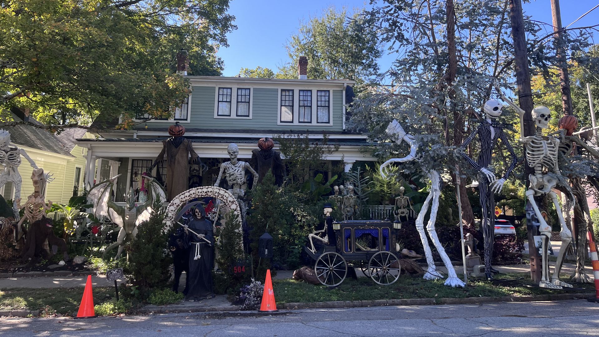 Front yard Halloween display on a light-green, two-story house: numerous white skeletons, pumpkins, and spooky props around a wagon; blue sky, trees, and overhead power lines.