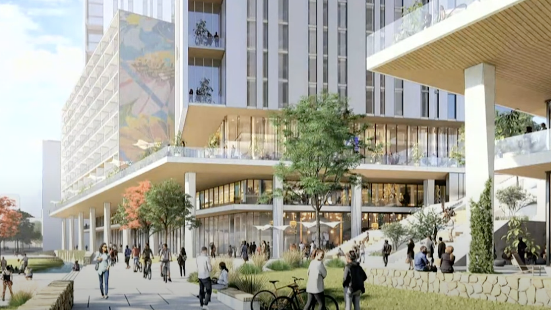 Modern building with large glass windows, balconies, people walking and biking nearby, green trees and outdoor seating areas on a sunny day in an urban plaza.