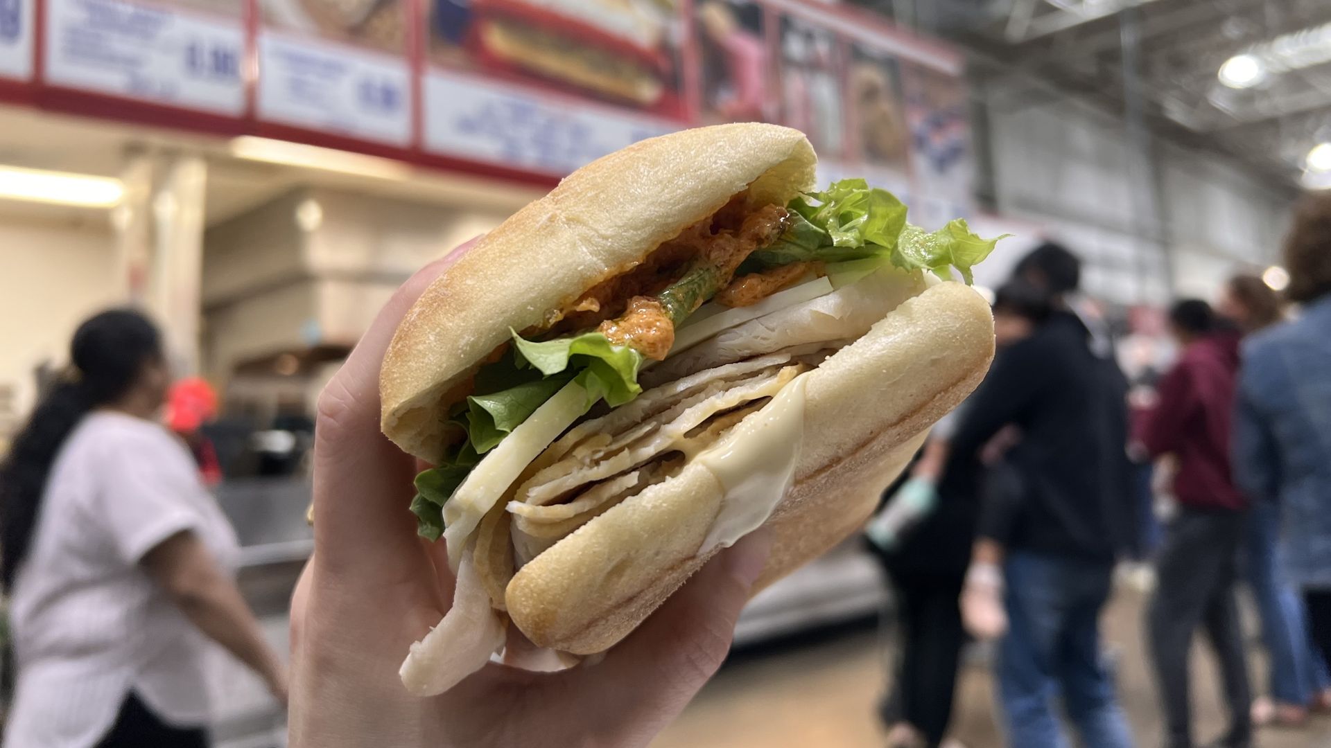 A picture of a ciabatta bread sandwich with lettuce, a sauce, turkey and cheese being held in the air by a hand while the background of people and a Costco menu are blurred
