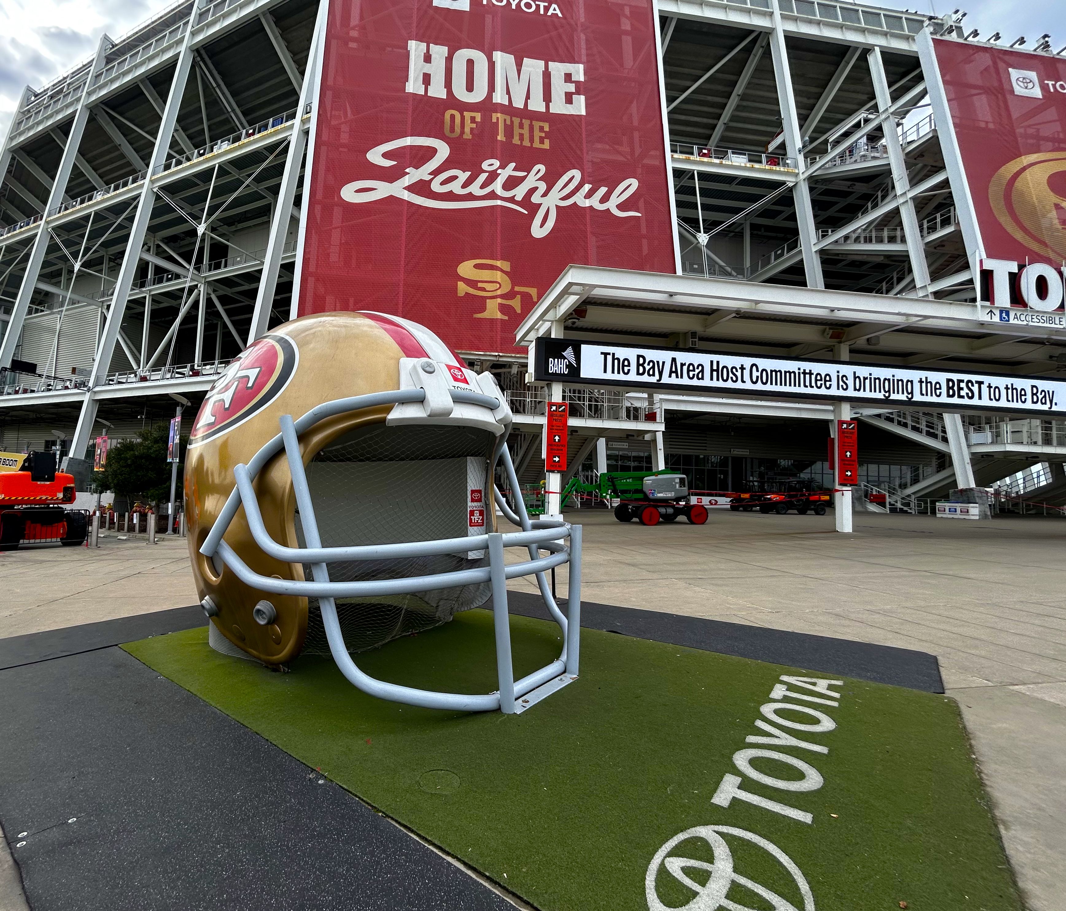 Large gold San Francisco 49ers helmet sculpture outside a stadium with red banners reading "Home of the Faithful" and a Toyota logo on green turf.