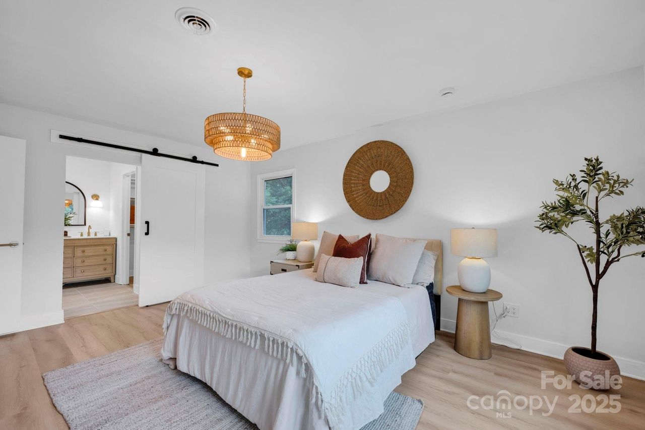 Bright modern bedroom with white walls, light wood floor, white bedding, circular woven wall decor, two nightstands with lamps, potted plant, and sliding door partially open to bathroom