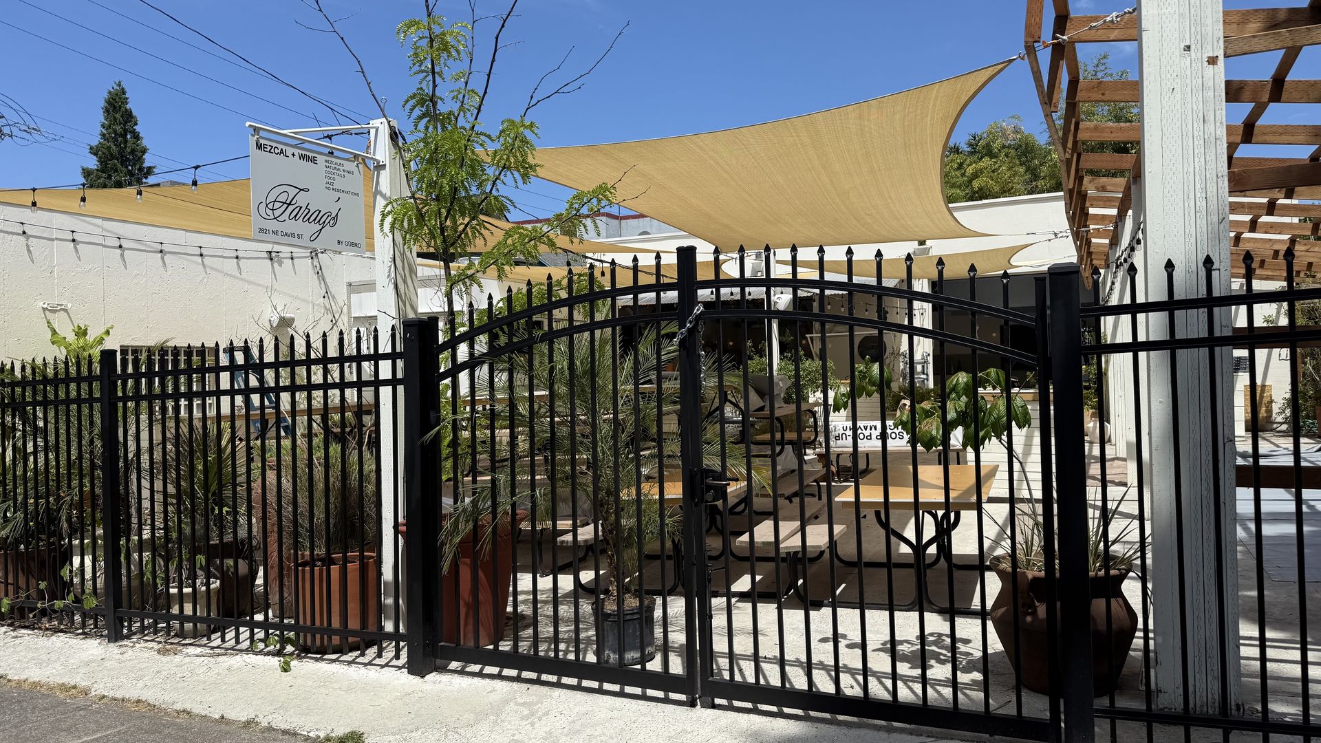 Outdoor seating area of a restaurant named Farag's Mezcal + Wine in Portland, Oregon, enclosed by a black iron fence with beige sunshades overhead and potted plants throughout the patio."